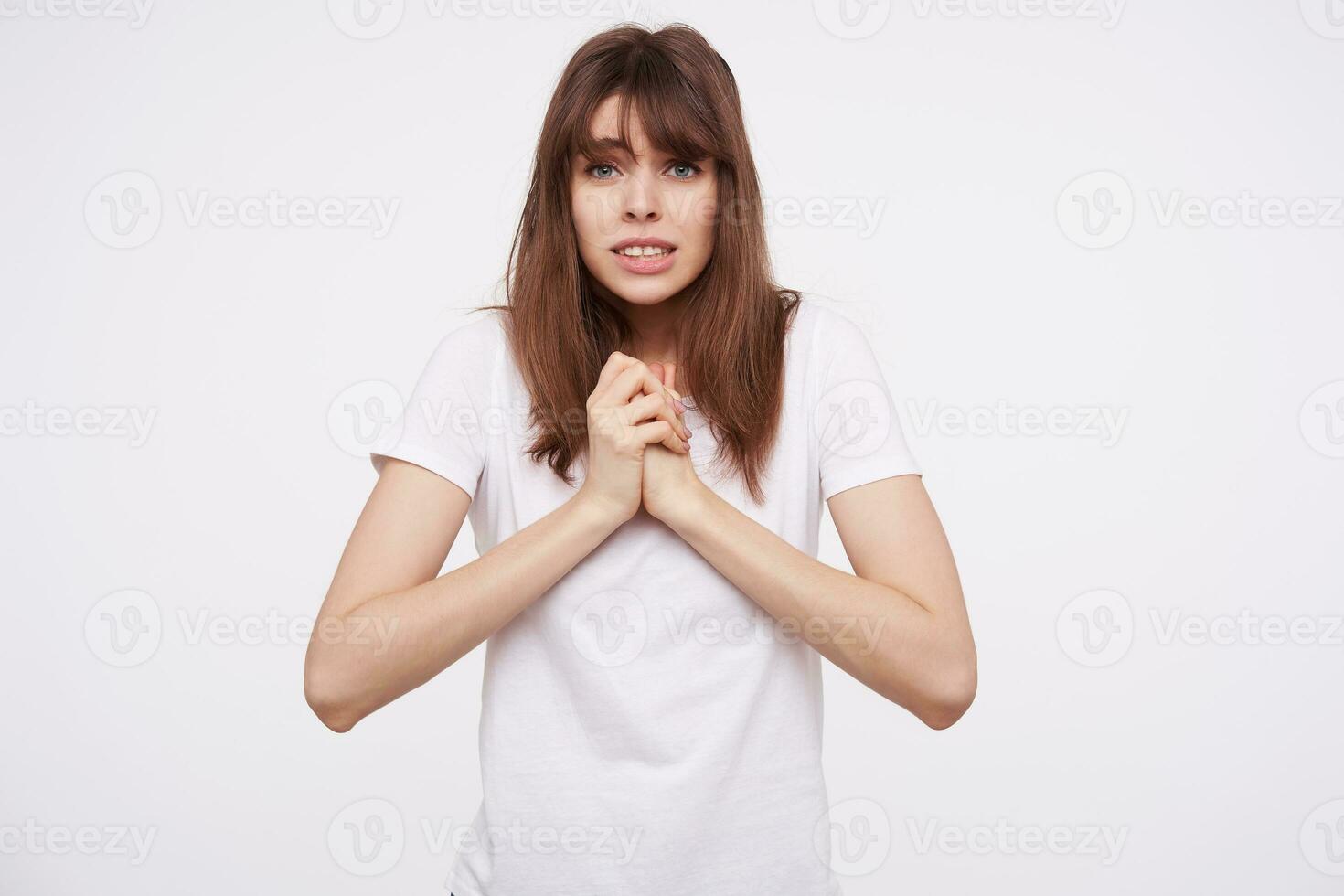 schot van bang jong brunette vrouw gekleed in wit eenvoudig t-shirt houden haar verheven handen samen en tonen haar tanden terwijl op zoek Bij camera met verbaasd gezicht, geïsoleerd over- wit achtergrond foto