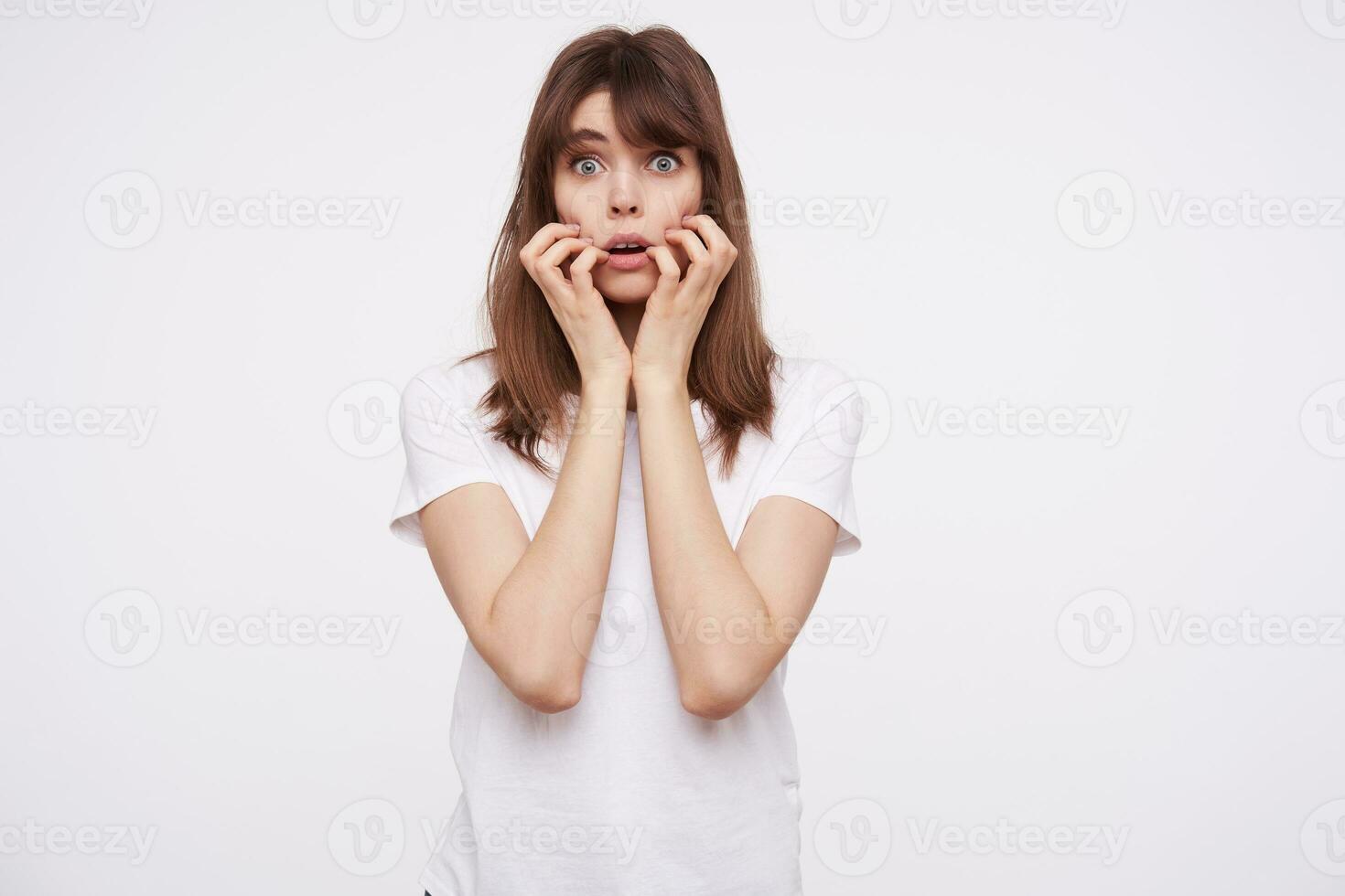 bang jong brunette vrouw met gewoontjes kapsel houden haar gezicht met verheven handen en afronding haar blauw ogen terwijl op zoek bang naar camera, geïsoleerd over- wit achtergrond foto