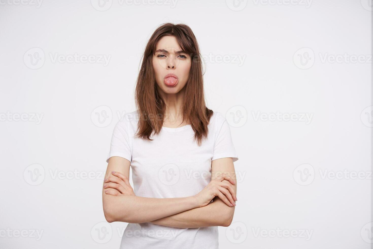 studio foto van jong mooi donker haren vrouw tonen tong Bij camera en vouwen handen Aan haar borst terwijl staand over- wit achtergrond in gewoontjes kleren