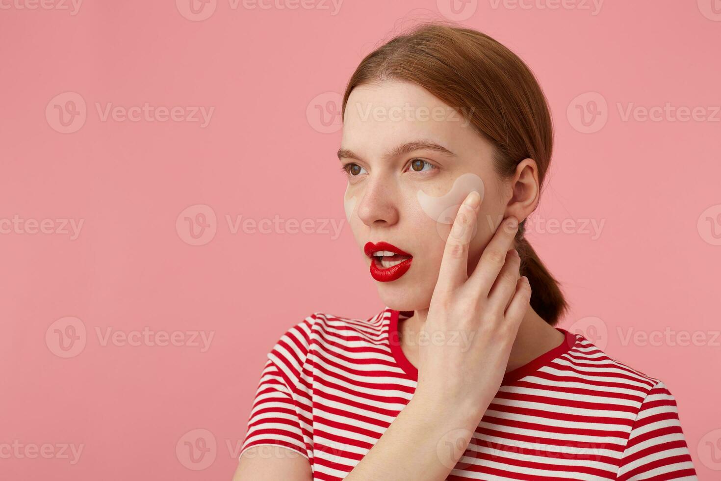 dichtbij omhoog van denken jong roodharig meisje met rood lippen en met patches onder de ogen, draagt in een rood gestreept t-shirt, looks weg, accenten de wang, staat over- roze achtergrond. foto