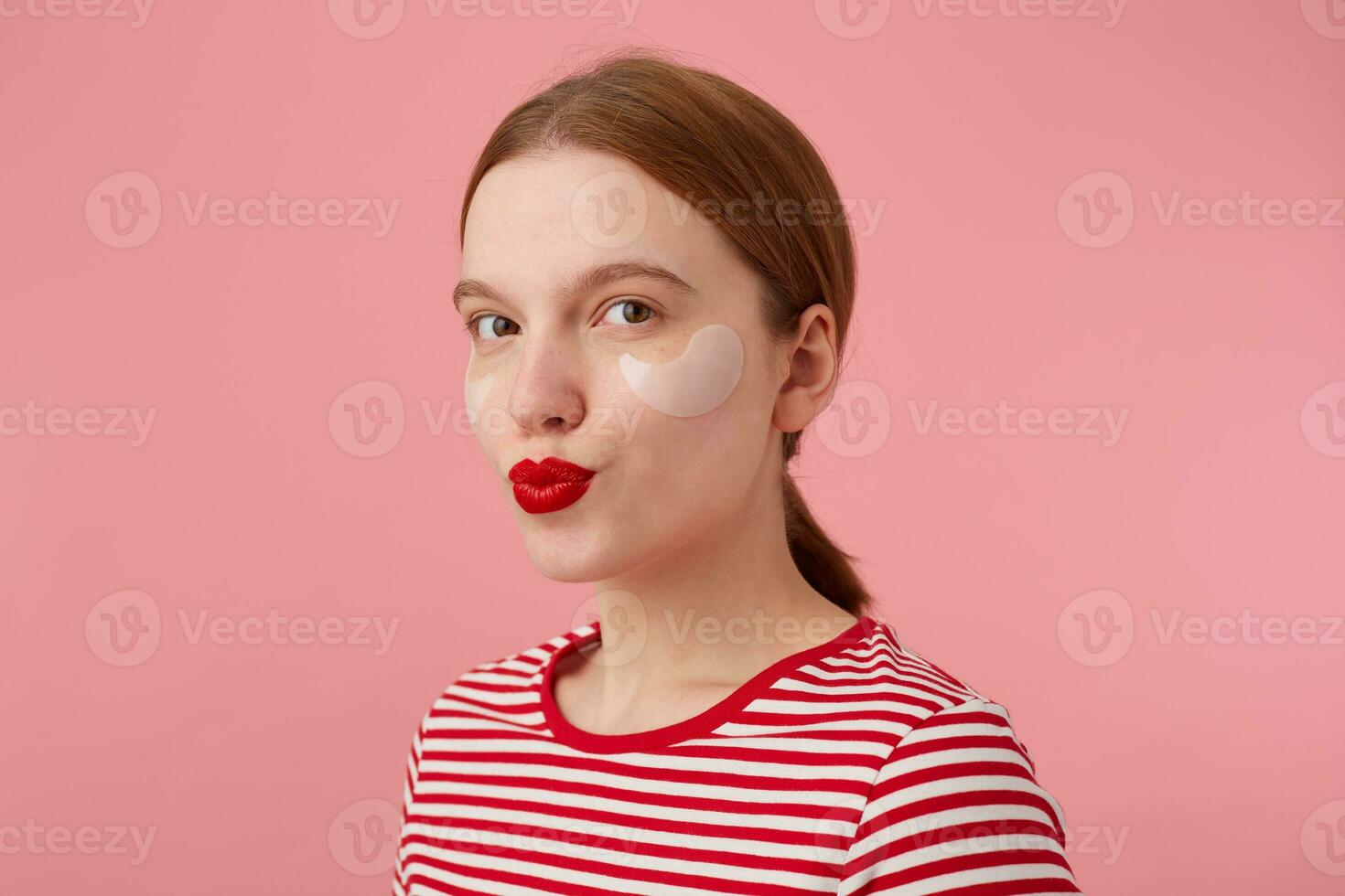 portret van schattig jong glimlachen roodharig meisje met rood lippen en met patches onder de ogen, draagt in een rood gestreept t-shirt, looks Bij de camera en stuurt kus, staat over- roze achtergrond. foto