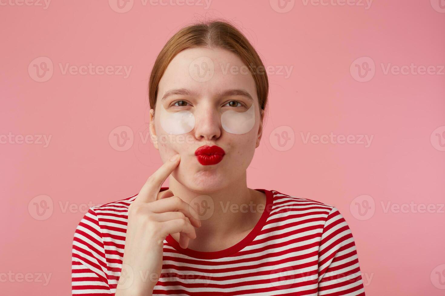 portret van denken jong roodharig meisje met rood lippen en met patches onder de ogen, draagt in een rood gestreept t-shirt, staat over- roze achtergrond, points naar zijn wang en opsluiten Bij de camera. foto