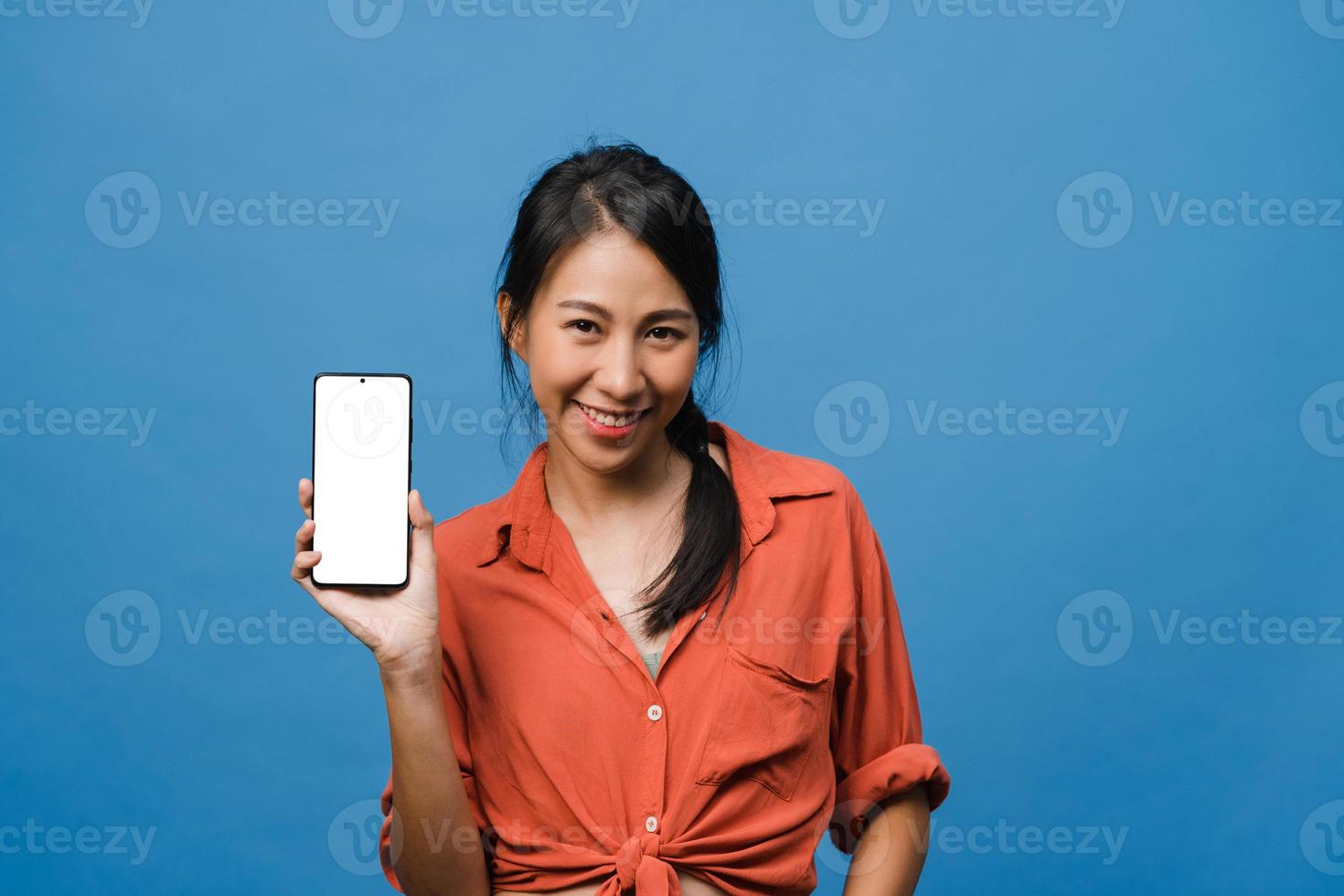 jonge aziatische dame toont een leeg smartphonescherm met positieve uitdrukking, glimlacht breed, gekleed in casual kleding en voelt zich gelukkig op een blauwe achtergrond. mobiele telefoon met wit scherm in vrouwelijke hand. foto