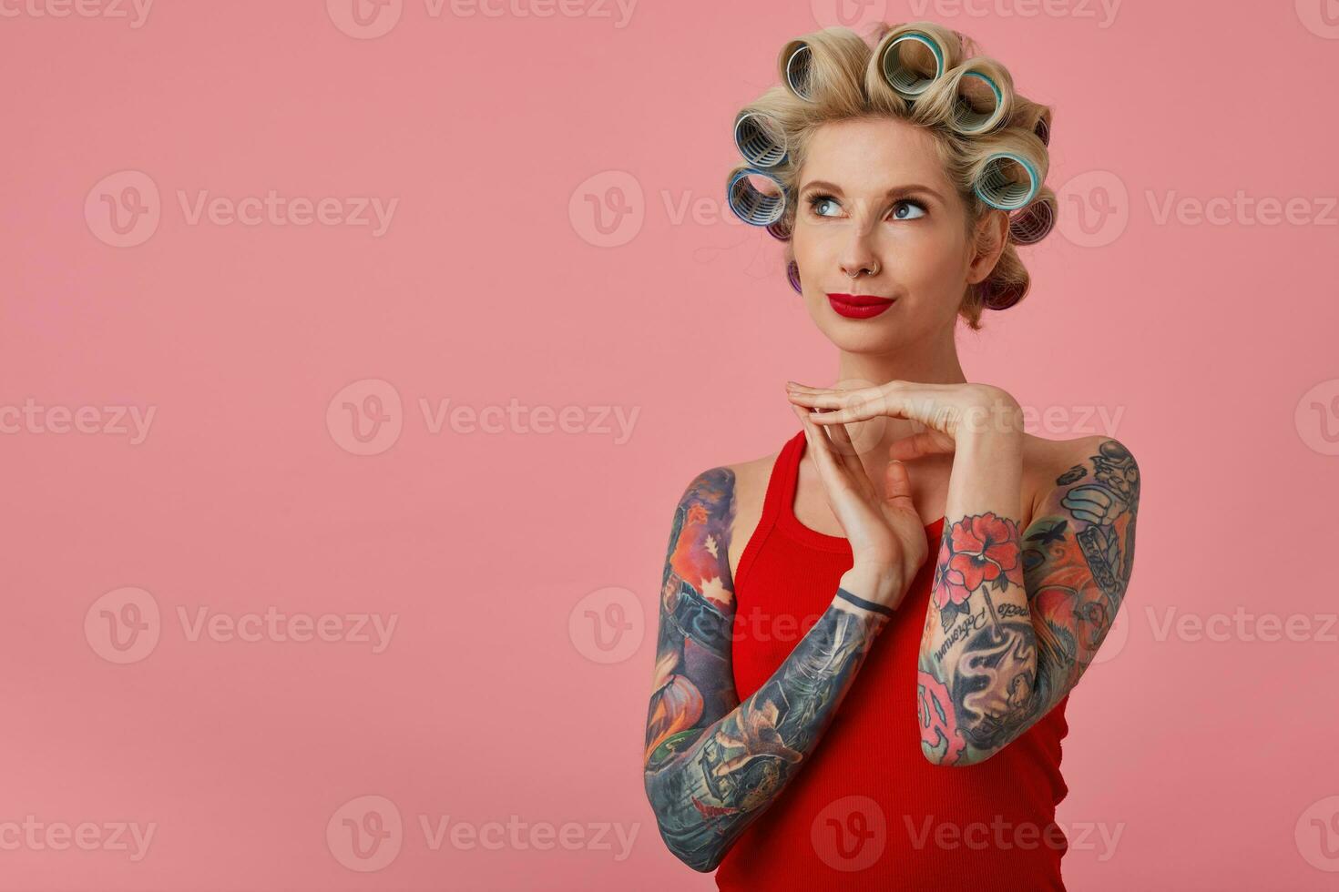 studio foto van jong charmant blond vrouw met tatoeages op zoek positief omhoog en vouwen verheven handen, hebben krulspelden Aan haar hoofd terwijl poseren over- roze achtergrond