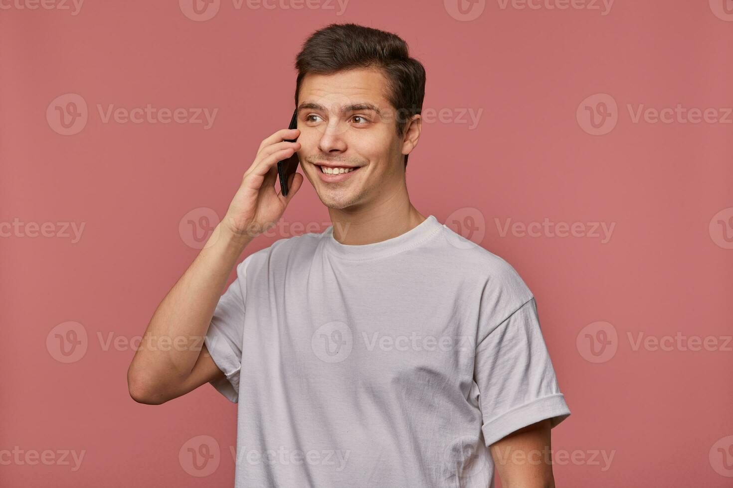 portret van knap jong bruin ogen Mens in grijs t-shirt op zoek terzijde met charmant oprecht glimlach, Holding mobiel telefoon in hand- en hebben aangenaam gesprek, staand over- roze achtergrond foto