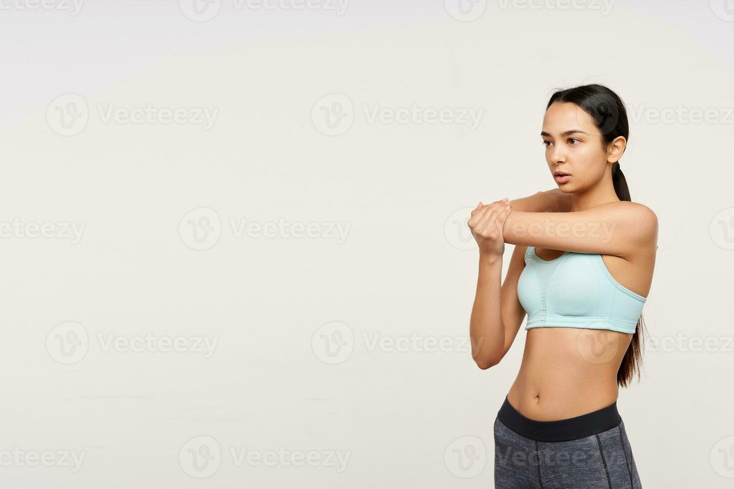 binnen- foto van jong slank bruin haren vrouw gekleed in munt top en grijs leggins uitrekken haar handen voordat training terwijl staand over- wit achtergrond