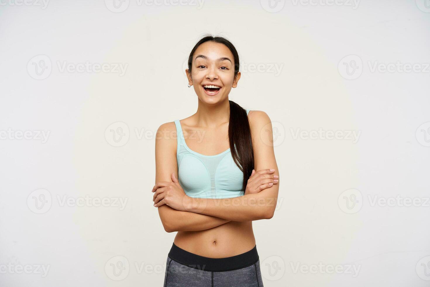 tiener- sportief meisje, gelukkig op zoek Aziatisch vrouw met donker lang haar. vervelend sportkleding en hebben een groot glimlach, houdt armen gekruiste Aan een borst. aan het kijken Bij de camera geïsoleerd over- wit achtergrond foto