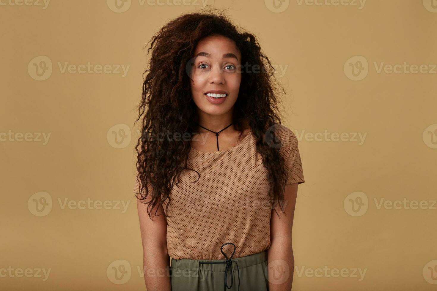 opgewonden jong lief bruin haren gekruld vrouw met donker huid verhogen verbaasd haar wenkbrauwen terwijl op zoek positief Bij camera, geïsoleerd over- beige achtergrond foto