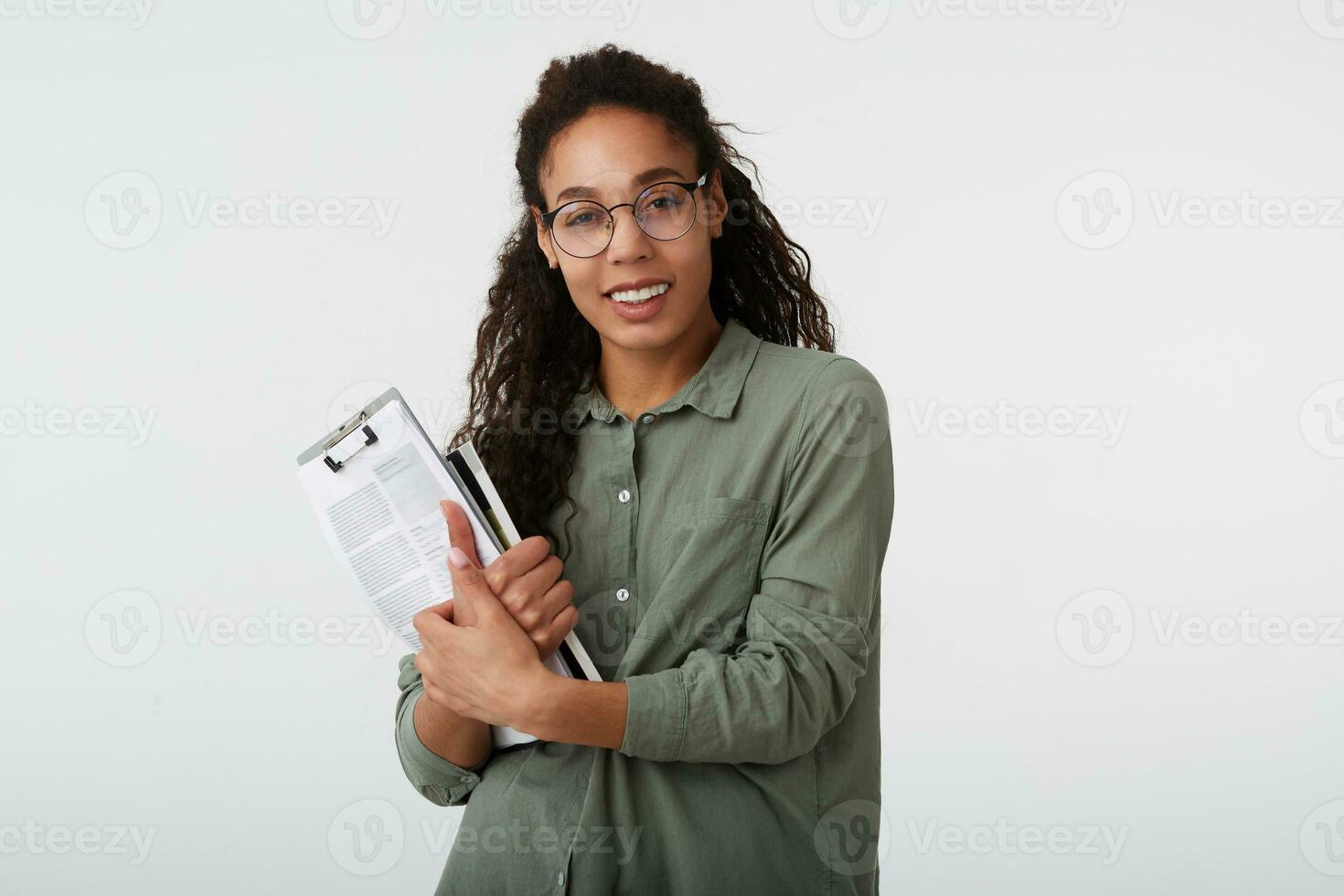 portret van charmant jong donker haren gekruld vrouw met donker huid houden leerboeken in haar handen en op zoek graag Bij camera met breed glimlach, geïsoleerd over- wit achtergrond foto