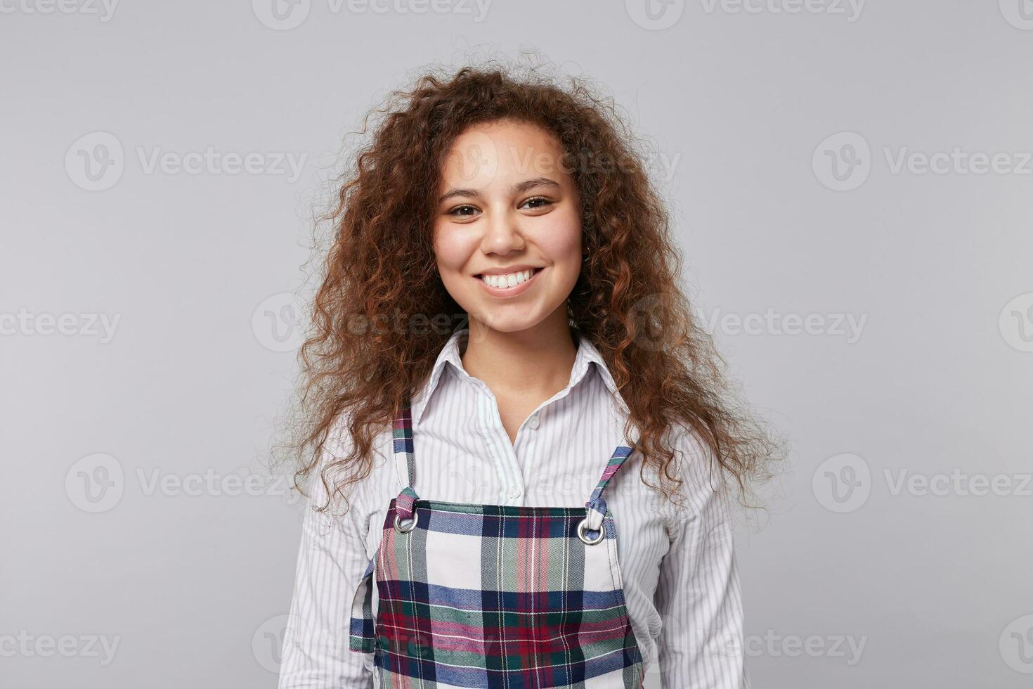 vrolijk jong lief lang haren gekruld brunette vrouw tonen haar wit perfect tanden terwijl glimlachen gelukkig Bij camera, staand over- grijs achtergrond foto