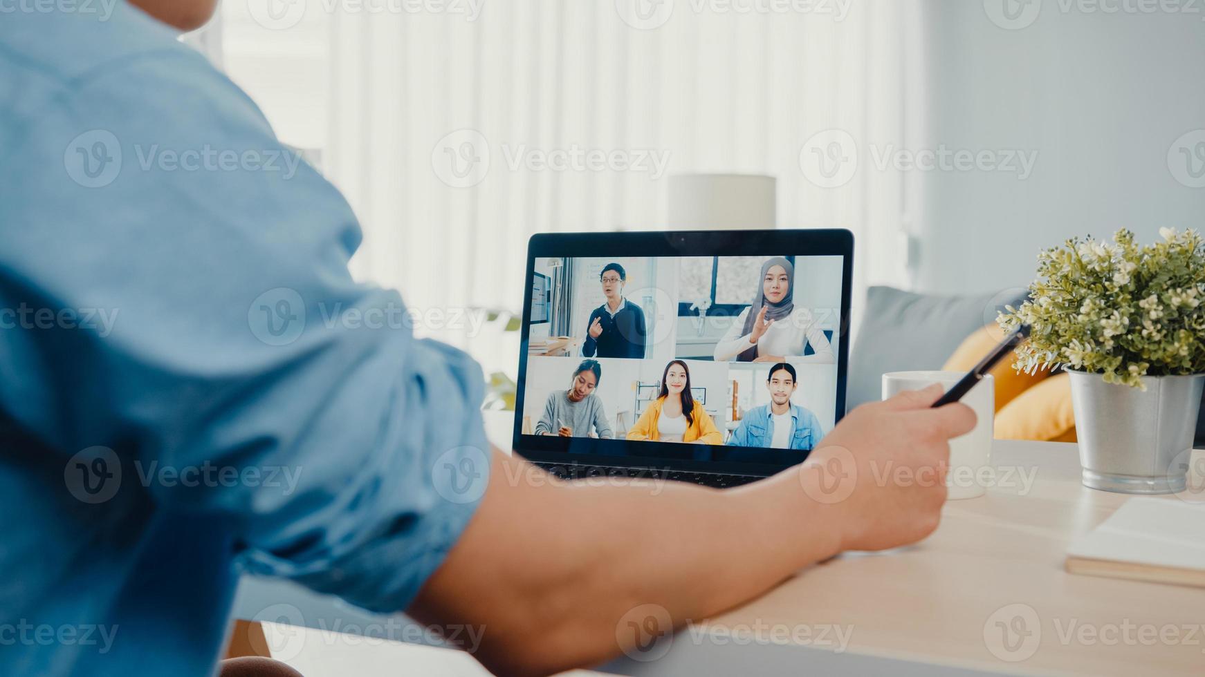 jonge Aziatische zakenman die laptop gebruikt, praat met collega's over het plan in een videogesprekvergadering terwijl hij vanuit huis in de woonkamer werkt. zelfisolatie, sociale afstand, quarantaine voor preventie van het coronavirus. foto