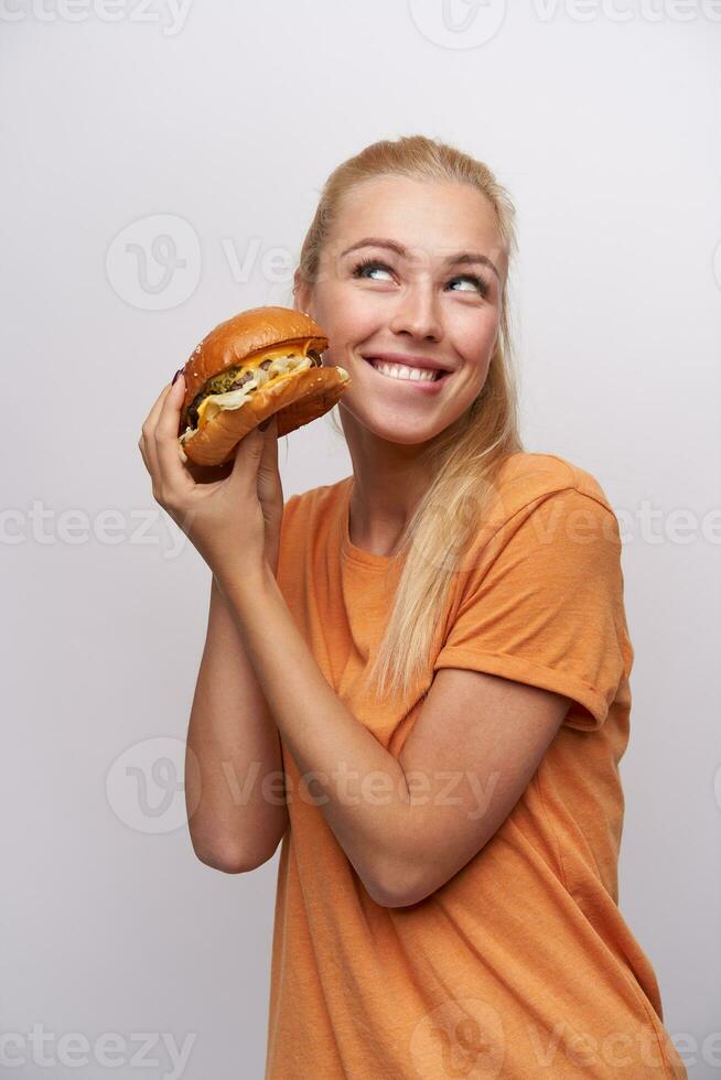 vrolijk jong mooi lang haren blond dame met gewoontjes kapsel verhogen handen met smakelijk hamburger en op zoek gelukkig terzijde met charmant glimlach, poseren over- wit achtergrond foto