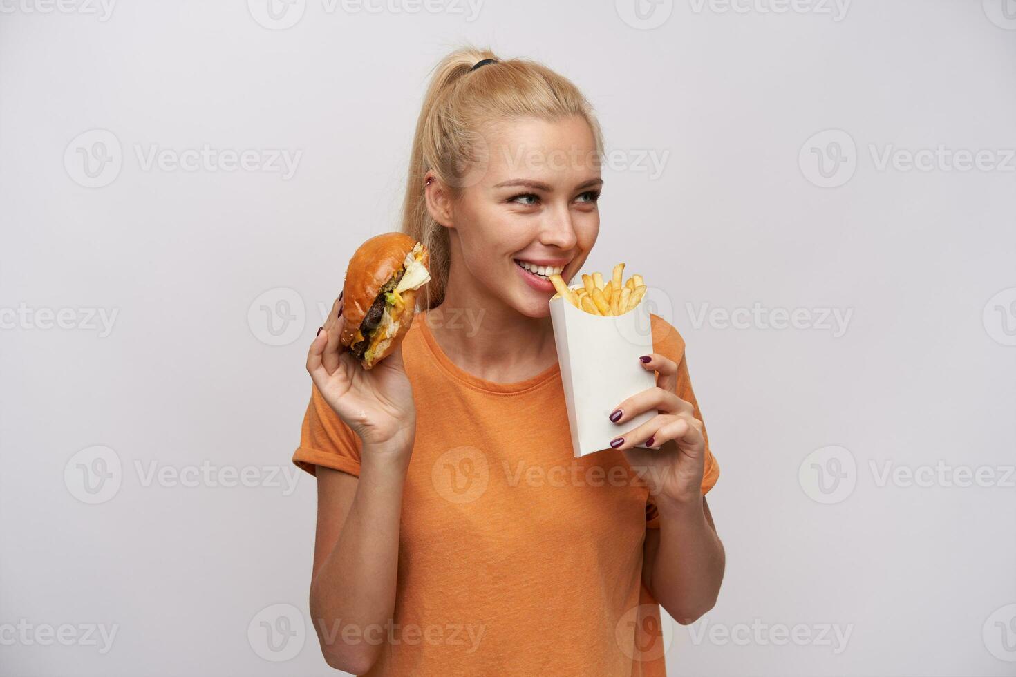 portret van lief jong blond vrouw met paardenstaart kapsel aan het eten Frans Patat en op zoek positief terzijde, glimlachen breed terwijl staand over- wit achtergrond foto