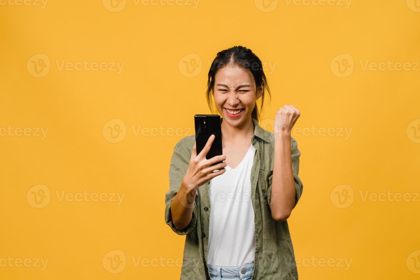 verraste jonge aziatische dame met behulp van mobiele telefoon met positieve uitdrukking, glimlacht breed, gekleed in casual kleding en staat geïsoleerd op gele achtergrond. gelukkige schattige blije vrouw verheugt zich over succes. foto