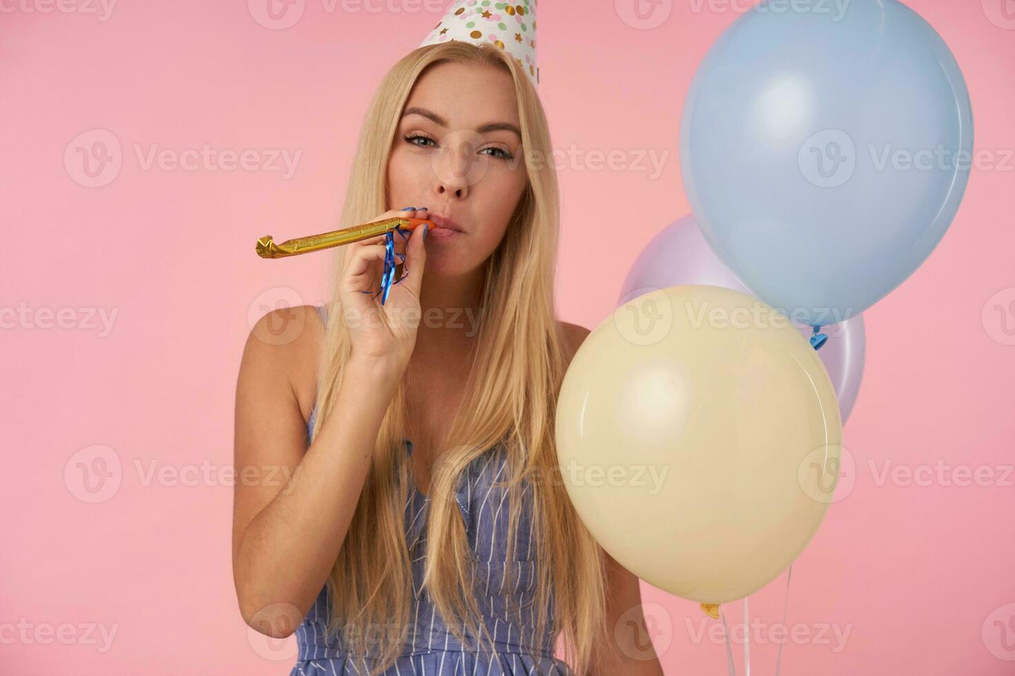 schot van mooi jong blond vrouw met lang haar- poseren in veelkleurig helium ballonnen, vieren vakantie, vervelend feestelijk kleren en ijshoorntje hoed, staand over- roze achtergrond met partij toeter foto