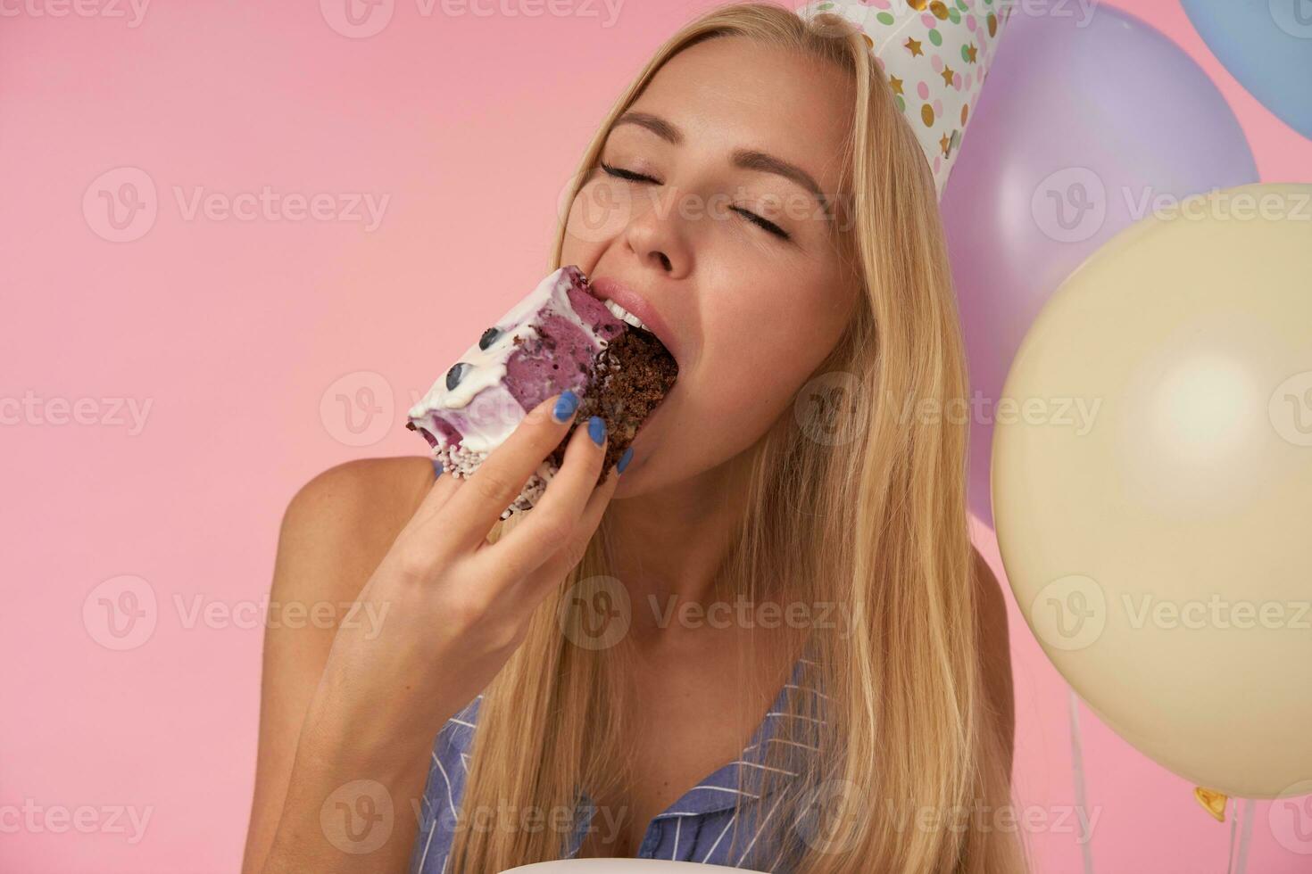 detailopname van mooi lang haren blond vrouw genieten van smakelijk verjaardag taart, houden ogen Gesloten terwijl tonen haar aangenaam emoties over- roze achtergrond, poseren in veelkleurig lucht ballonnen foto