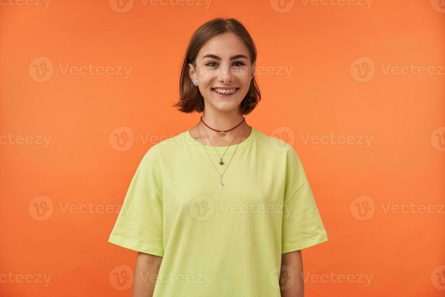 jong mooi vrouw met kort haar, breed glimlach, gelukkig op zoek. aan het kijken Bij de camera geïsoleerd over- oranje achtergrond. vervelend groen t-shirt, tanden een beugel en ketting. foto