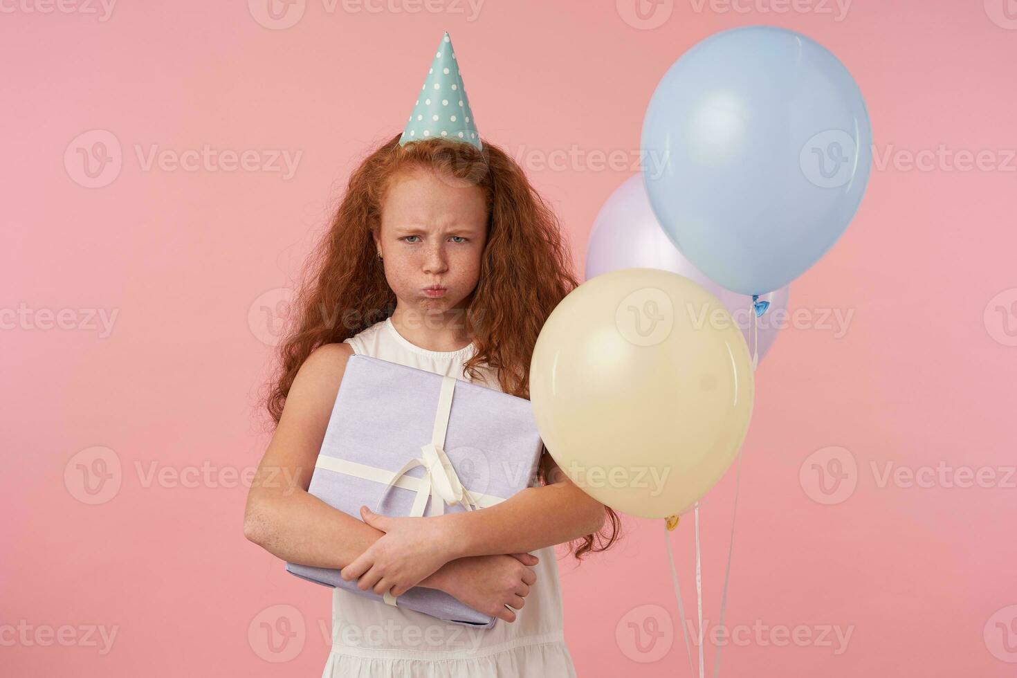 beledigd weinig meisje met lang foxy haar- Holding verpakt Cadeau doos, op zoek in camera helaas en pruilen, geïsoleerd over- roze achtergrond met gekleurde lucht ballonnen, vervelend wit elegant jurk foto