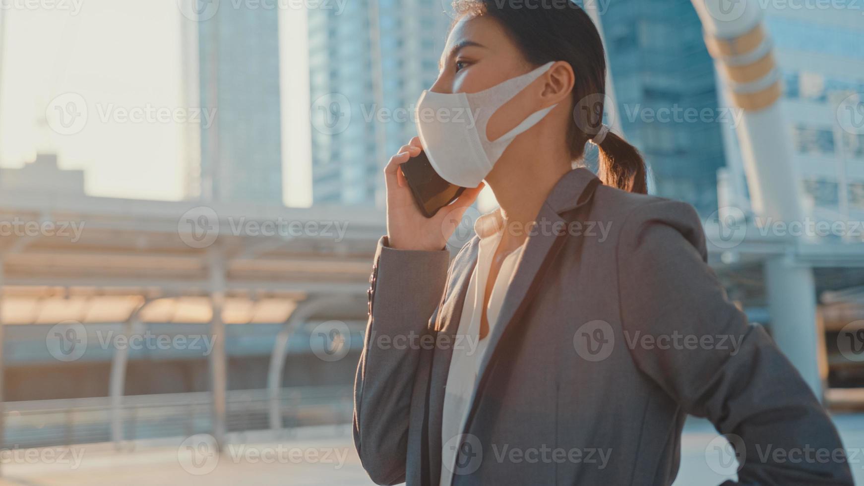 jonge Aziatische zakenvrouw in mode-kantoorkleding draagt een medisch gezichtsmasker, praat via de telefoon terwijl ze alleen buiten in de stedelijke stad loopt. zaken onderweg, sociale afstand om verspreiding van het covid-19-concept te voorkomen. foto
