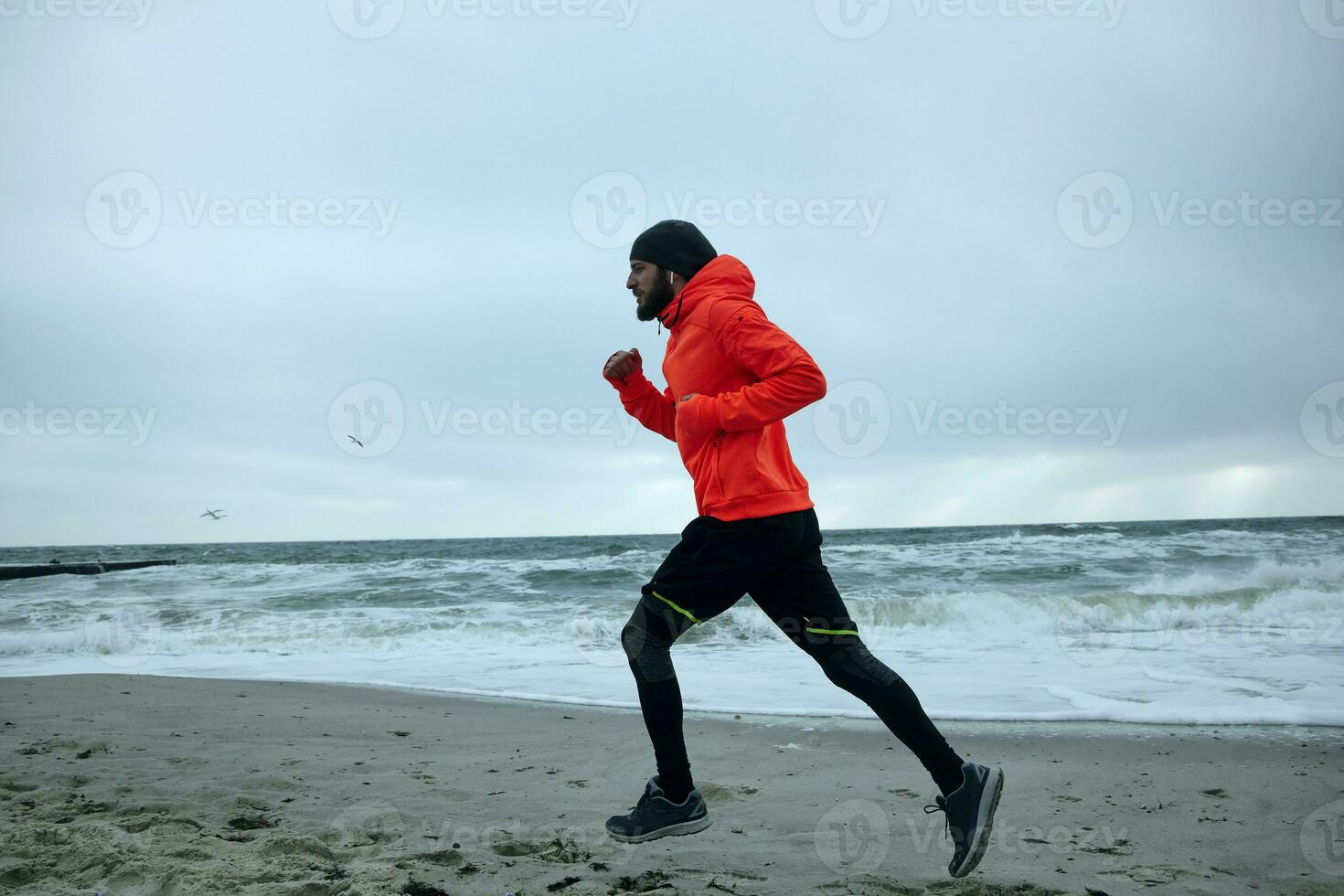 beeld van jong knap gebaard sportman luisteren naar favoriete muziek- lijst terwijl werken uit en rennen door kust Aan vroeg bewolkt ochtend. geschiktheid en sport concept foto