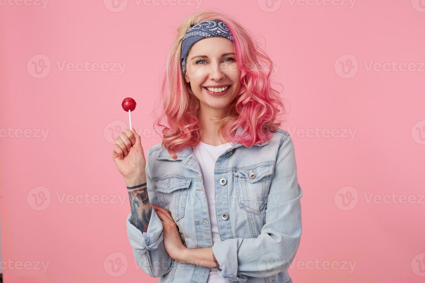 jong positief mooi roze haren dame in denim sjir, Holding een lolly, looks Bij de camera en breed lachend, staat over- roze achtergrond. foto