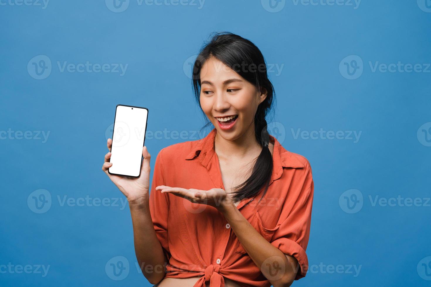 jonge aziatische dame toont een leeg smartphonescherm met positieve uitdrukking, glimlacht breed, gekleed in casual kleding en voelt zich gelukkig op een blauwe achtergrond. mobiele telefoon met wit scherm in vrouwelijke hand. foto