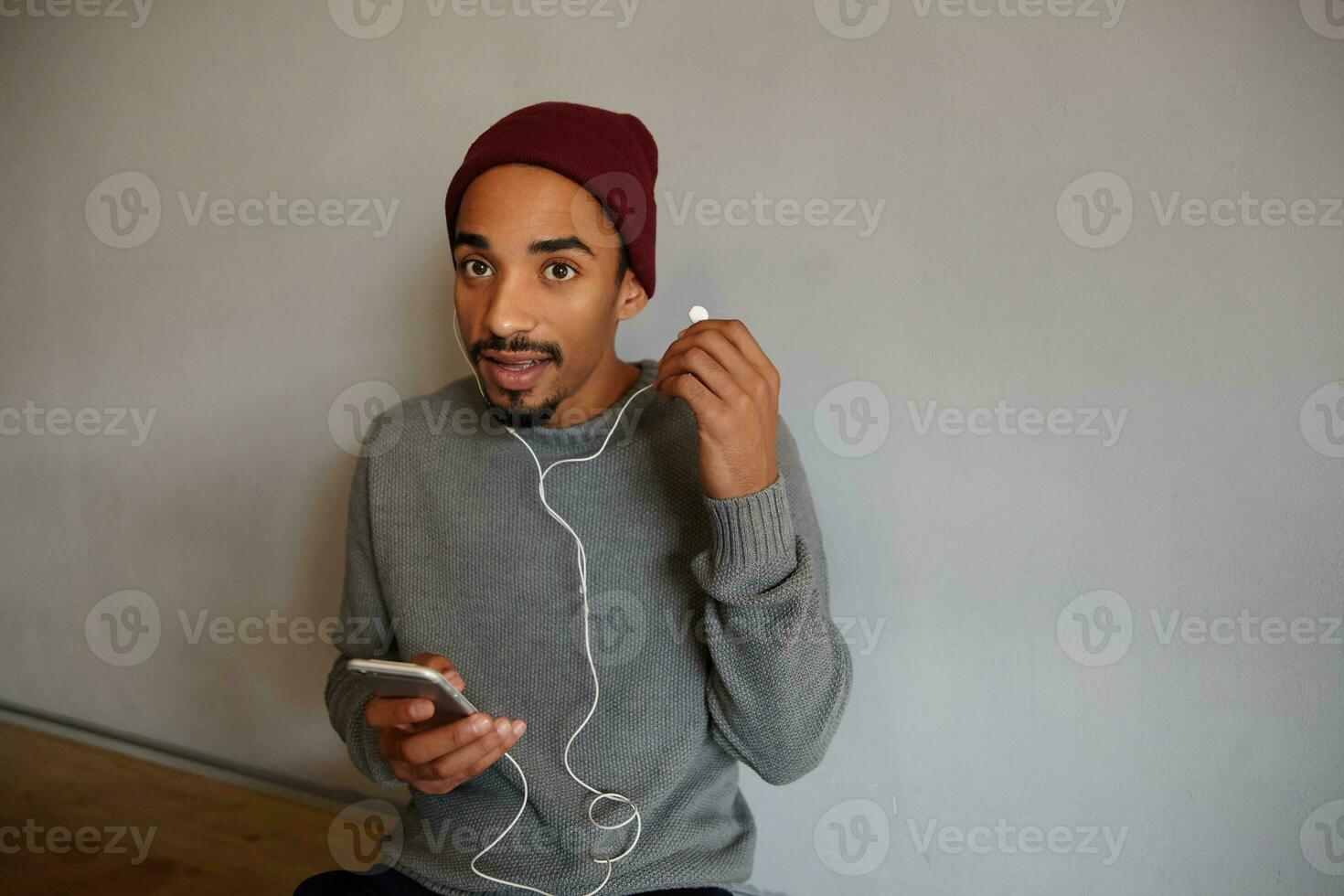 horizontaal schot van verbaasd met open ogen mooi jong donker huid mannetje nemen uit oortje en op zoek Bij camera verwonderd, gekleed in grijs trui en bordeaux hoofdtooi foto