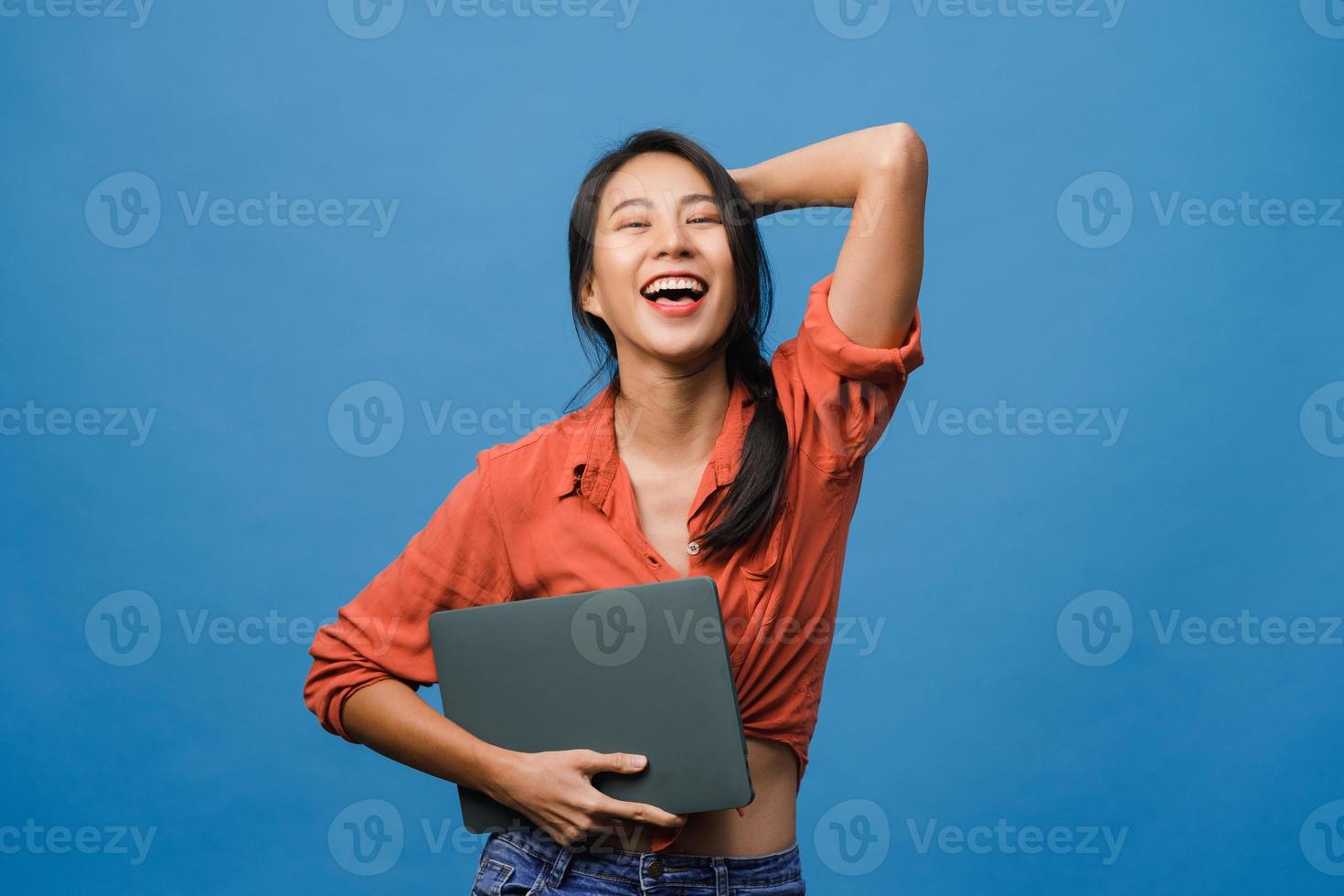 verraste jonge aziatische dame houdt laptop vast met positieve uitdrukking, glimlach breed, gekleed in casual kleding en kijkt naar camera op blauwe achtergrond. gelukkige schattige blije vrouw verheugt zich over succes. foto