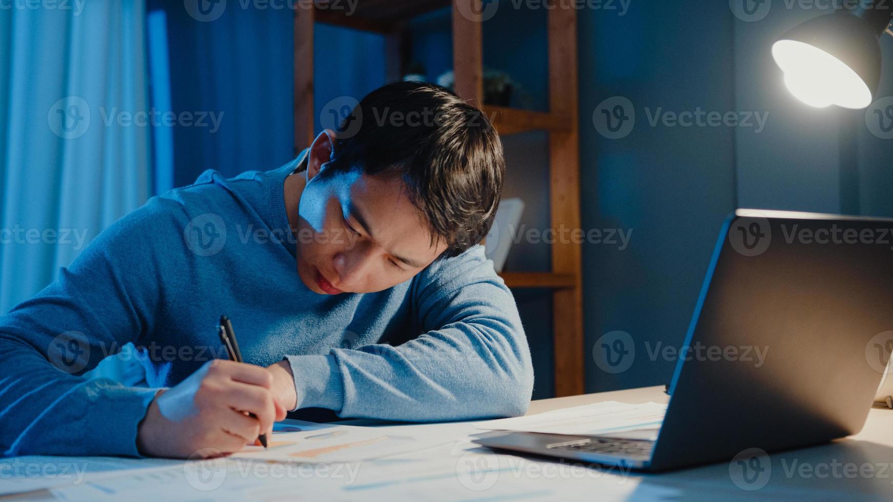 Aziatische freelance zakenman focus werktype op laptopcomputer bezig met vol papierwerk grafiek op bureau in woonkamer thuis overuren 's nachts, werk vanuit huis tijdens covid-19 pandemie concept. foto