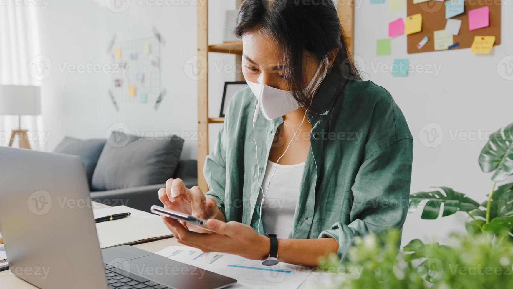 freelancer azië vrouwen dragen gezichtsmasker met smartphone die online via de website winkelt terwijl ze aan een bureau in de woonkamer zitten. thuiswerken, werken op afstand, sociale afstand, quarantaine voor coronavirus. foto