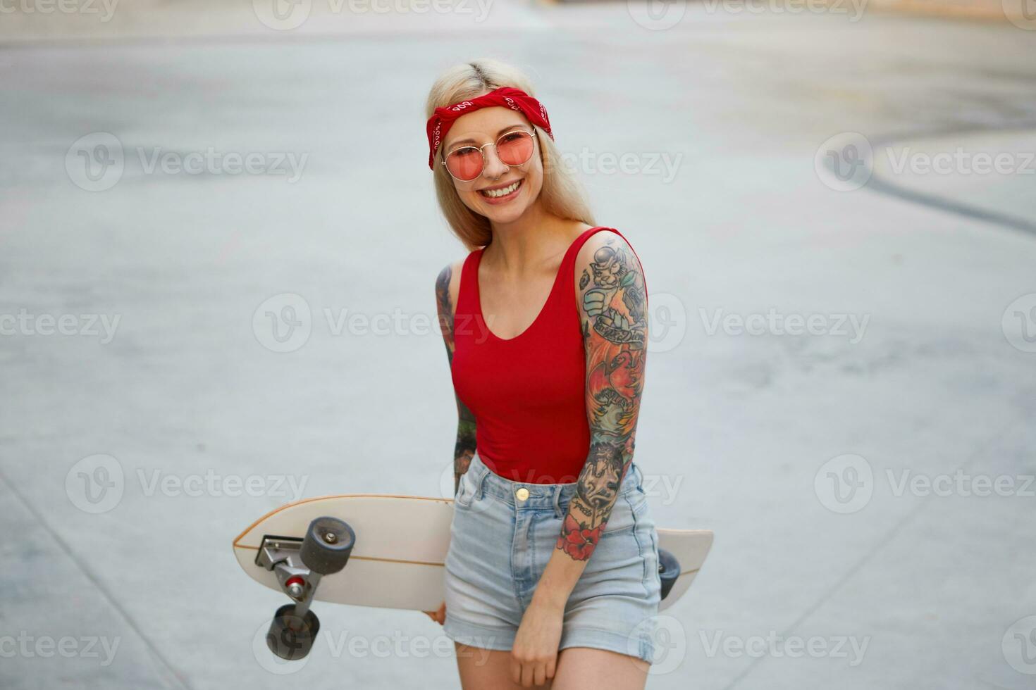 schattig blond dame met getatoeëerd armen in rood bril, rood t-shirt en denim shorts met een gebreid bandana Aan hoofd, Holding een longboard in hand, breed lachend, besteedt tijd in vleet park en geniet. foto