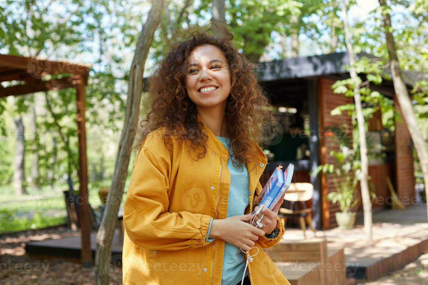 jong aantrekkelijk donker huid gekruld leerling dame Aan een cafe terras op zoek weg, vervelend in geel jas, Holding studieboeken, glimlacht breed, geniet de het weer. foto