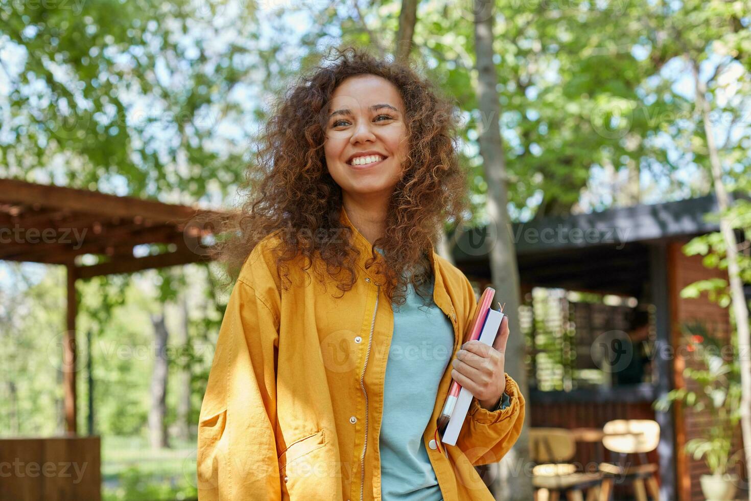 portret van glimlachen jong mooi donker huid gekruld leerling meisje Aan een cafe terras, Holding studieboeken, vervelend in geel jas, geniet de het weer. foto