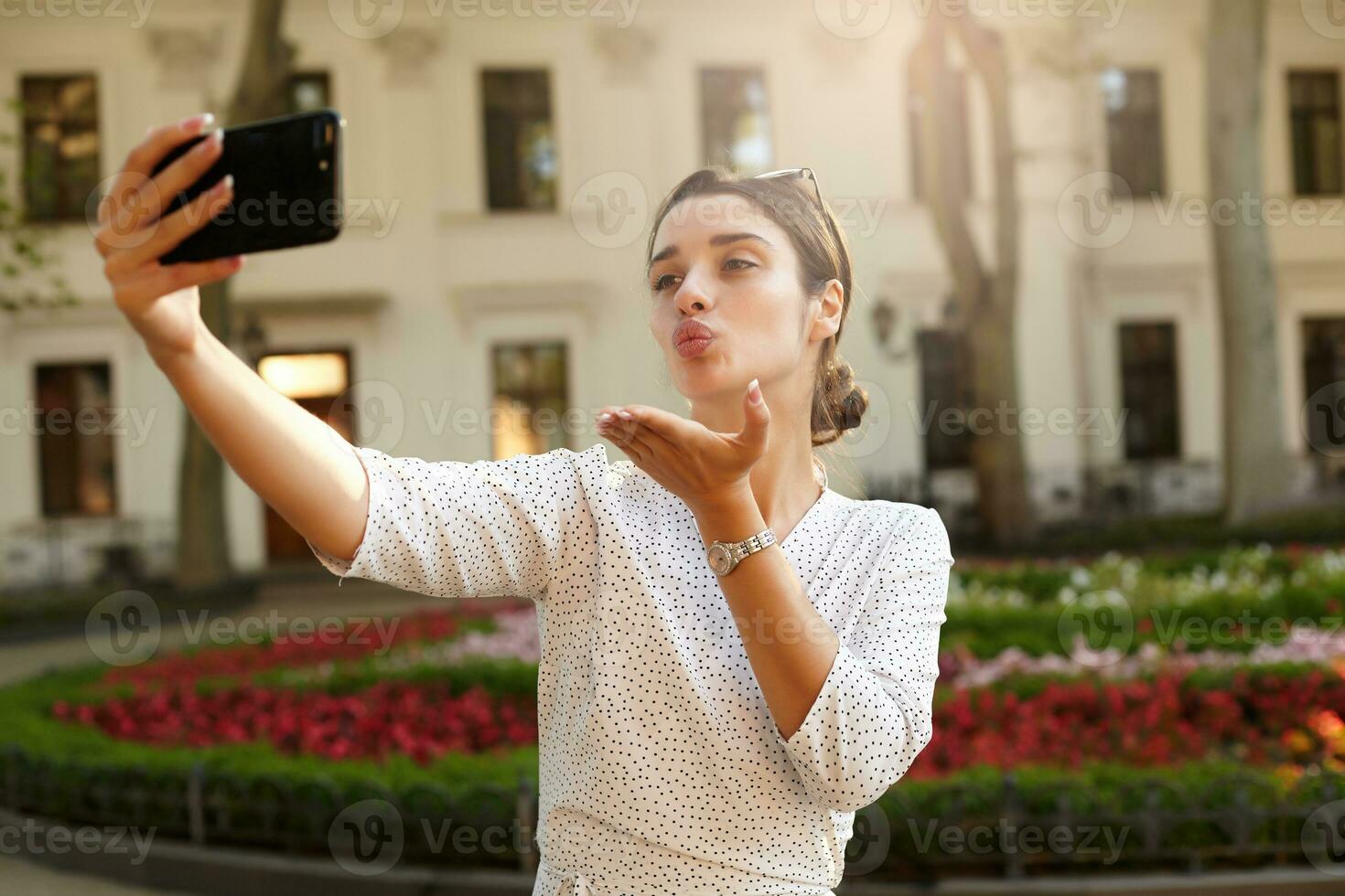 mooi jong donker haren vrouw met gewoontjes kapsel poseren buitenshuis Aan warm voorjaar dag met mobiel telefoon in verheven hand, blazen lucht kus naar camera terwijl maken foto van haarzelf