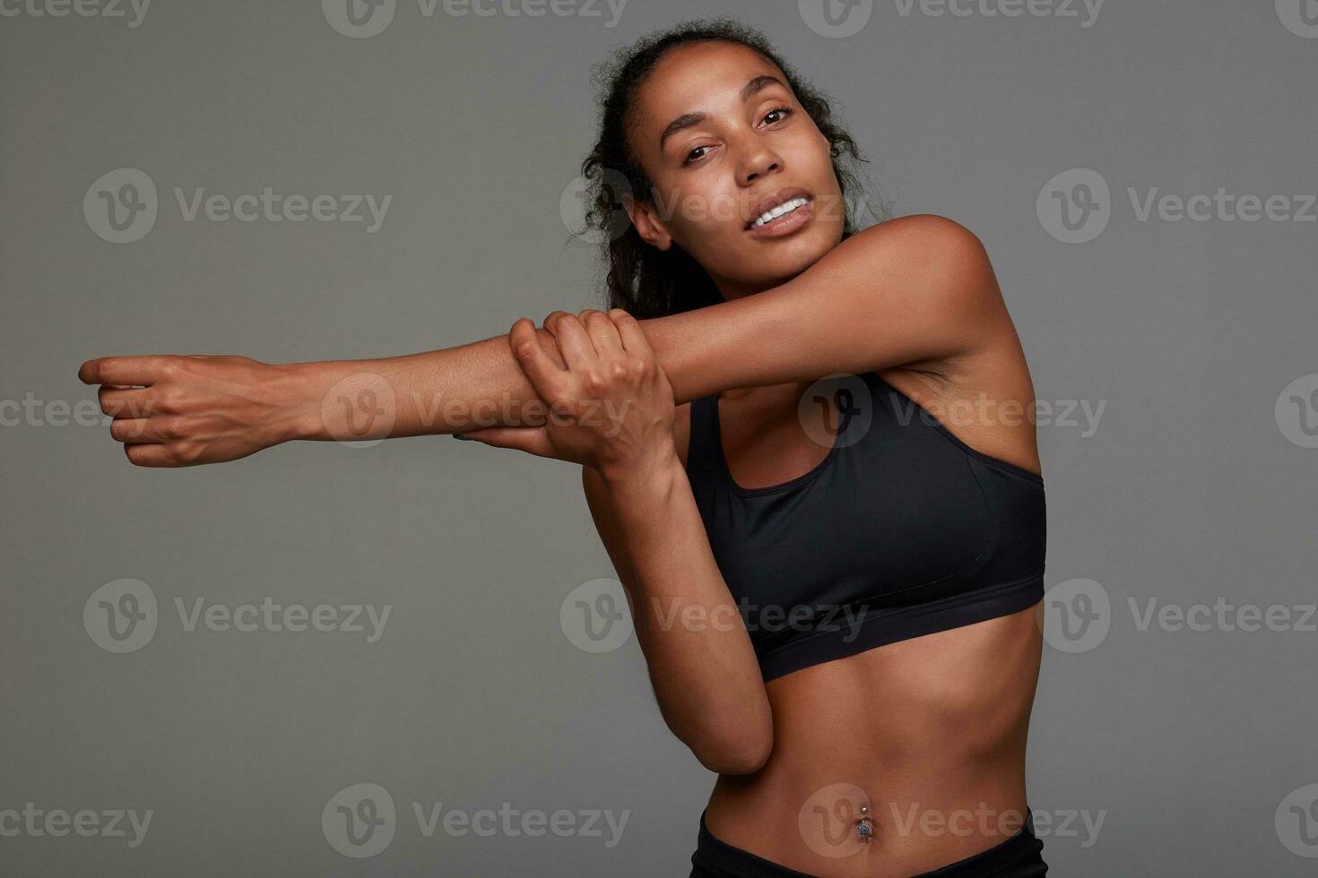 portret van sportief jong gekruld brunette vrouw met donker huid uitrekken haar handen voordat opleiding in Sportschool, gekleed in zwart atletisch slijtage terwijl poseren over- grijs achtergrond foto