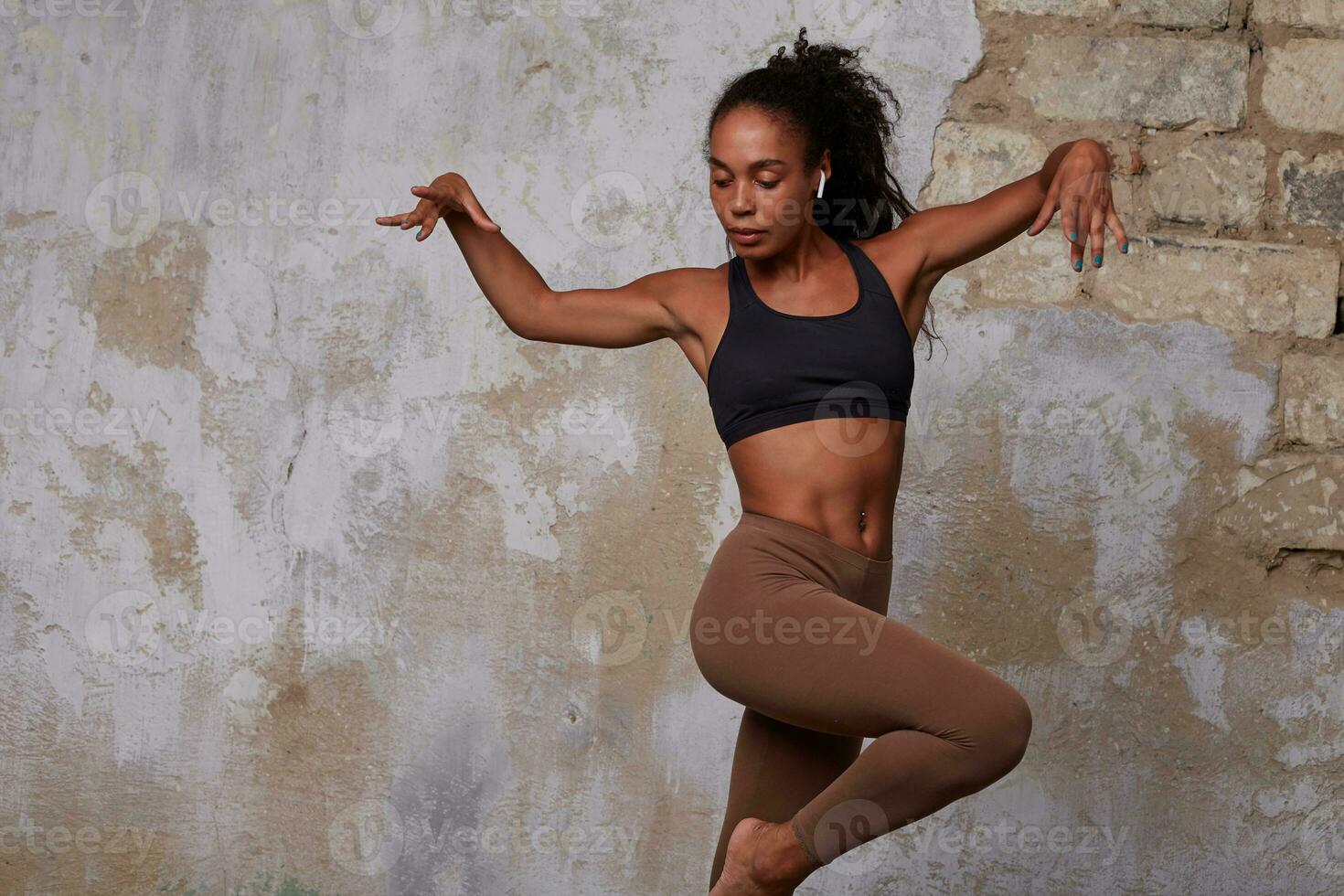 studio foto van jong slank donker huid gekruld brunette vrouw in zwart top en beige leggins voorbereidingen treffen modern dans terwijl poseren over- steen muur achtergrond