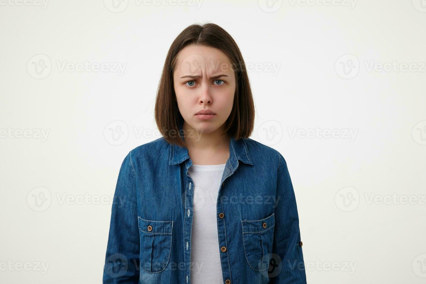 portret van jong van streek blauwe ogen brunette vrouw fronsen haar wenkbrauwen en houden lippen gevouwen terwijl op zoek Bij camera helaas, staand over- wit achtergrond foto