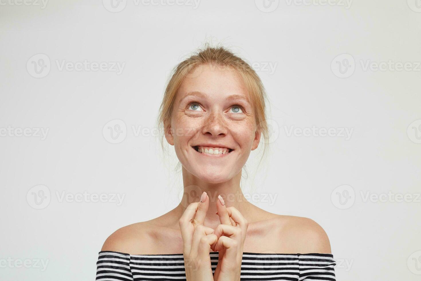 positief jong aantrekkelijk roodharige vrouw met gewoontjes kapsel glimlachen vrolijk terwijl op zoek dromerig omhoog, houden verheven handen onder haar kin terwijl staand over- wit achtergrond foto