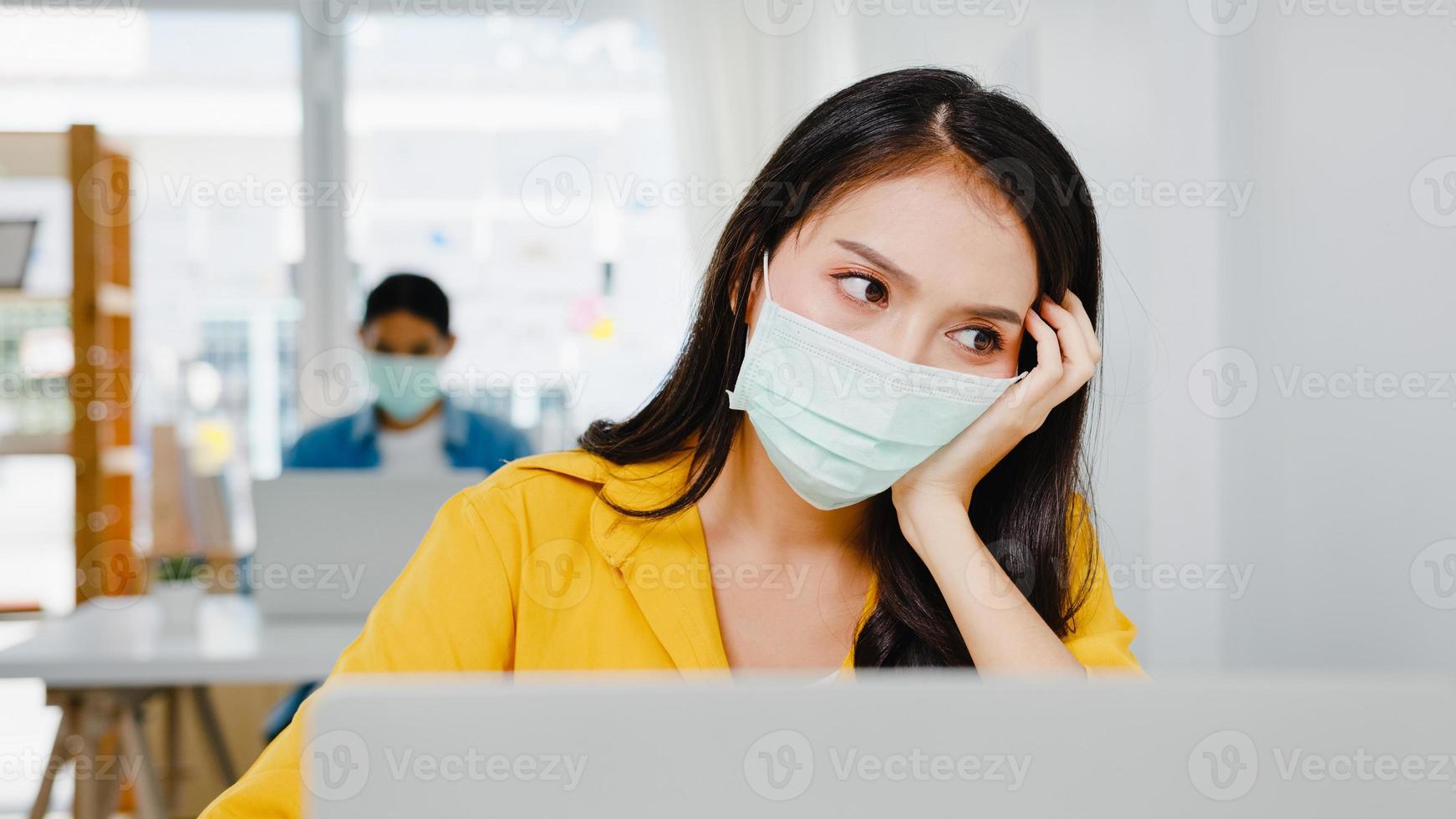 freelance azische vrouwen dragen gezichtsmasker met behulp van laptop hard werken op een nieuw normaal thuiskantoor. werken vanuit huis overbelasting, zelfisolatie, sociale afstand, quarantaine voor preventie van het coronavirus. foto