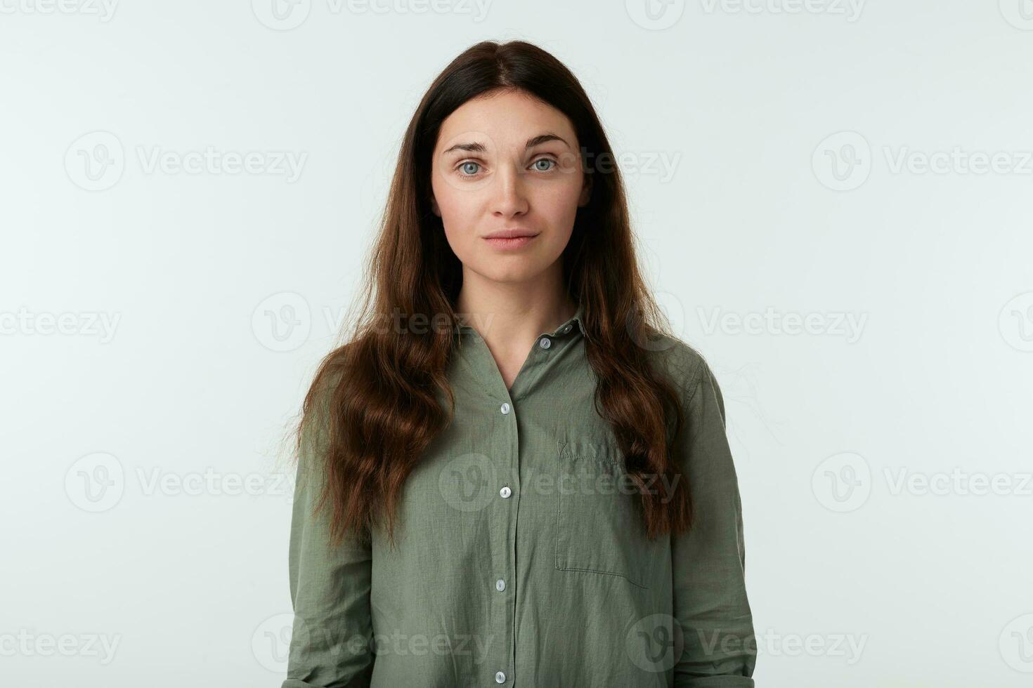 aantrekkelijk jong groene ogen vrouw met bruin golvend haar- op zoek rustig Bij camera met licht glimlach terwijl staand over- wit achtergrond in gewoontjes leger overhemd foto