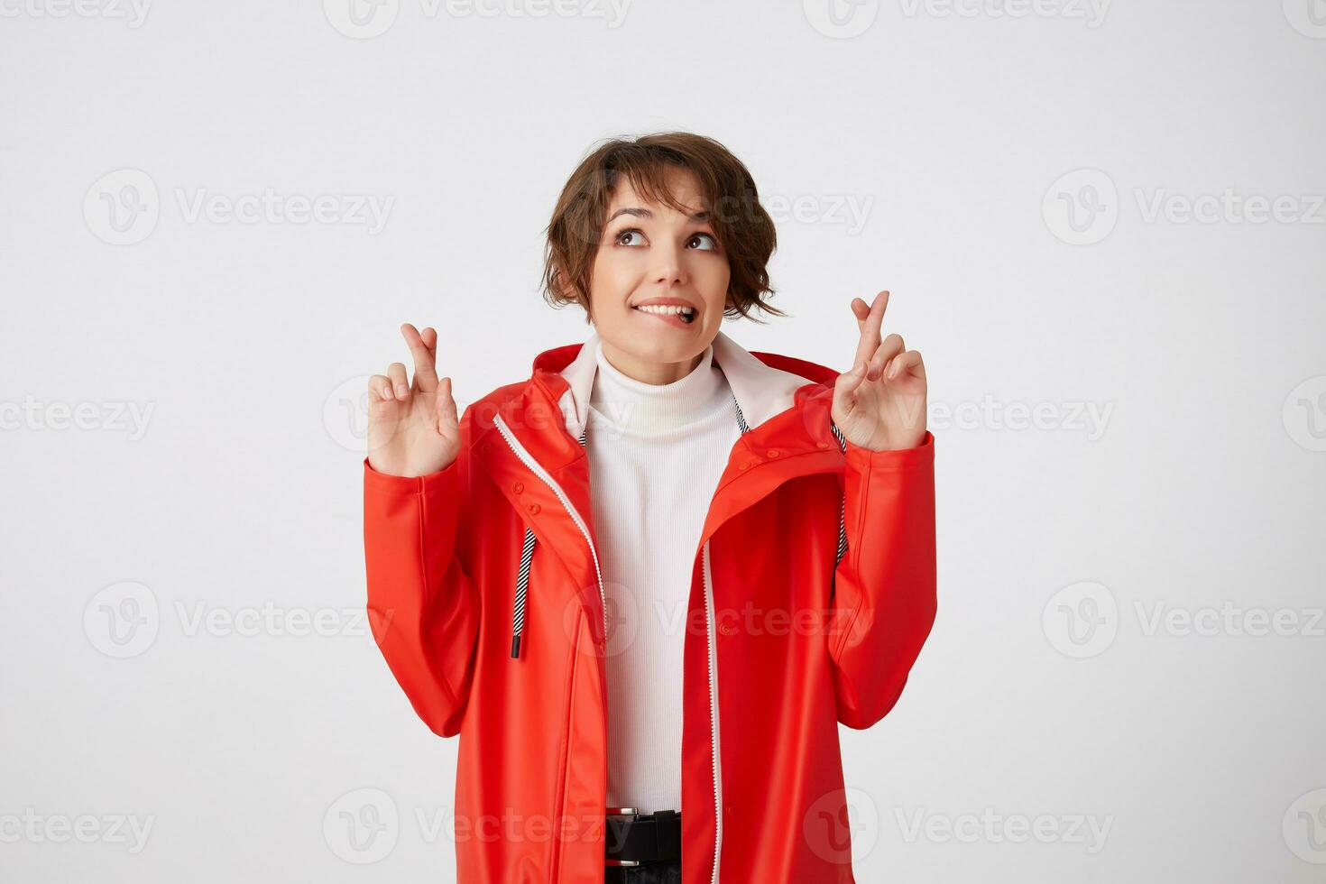 mooi hoor positief jong kort haren dame in wit golf en rood regen jas, bijt lippen, looks omhoog Bij de kopiëren ruimte, gekruiste vingers en hoopt voor geluk. staand over- wit achtergrond. foto