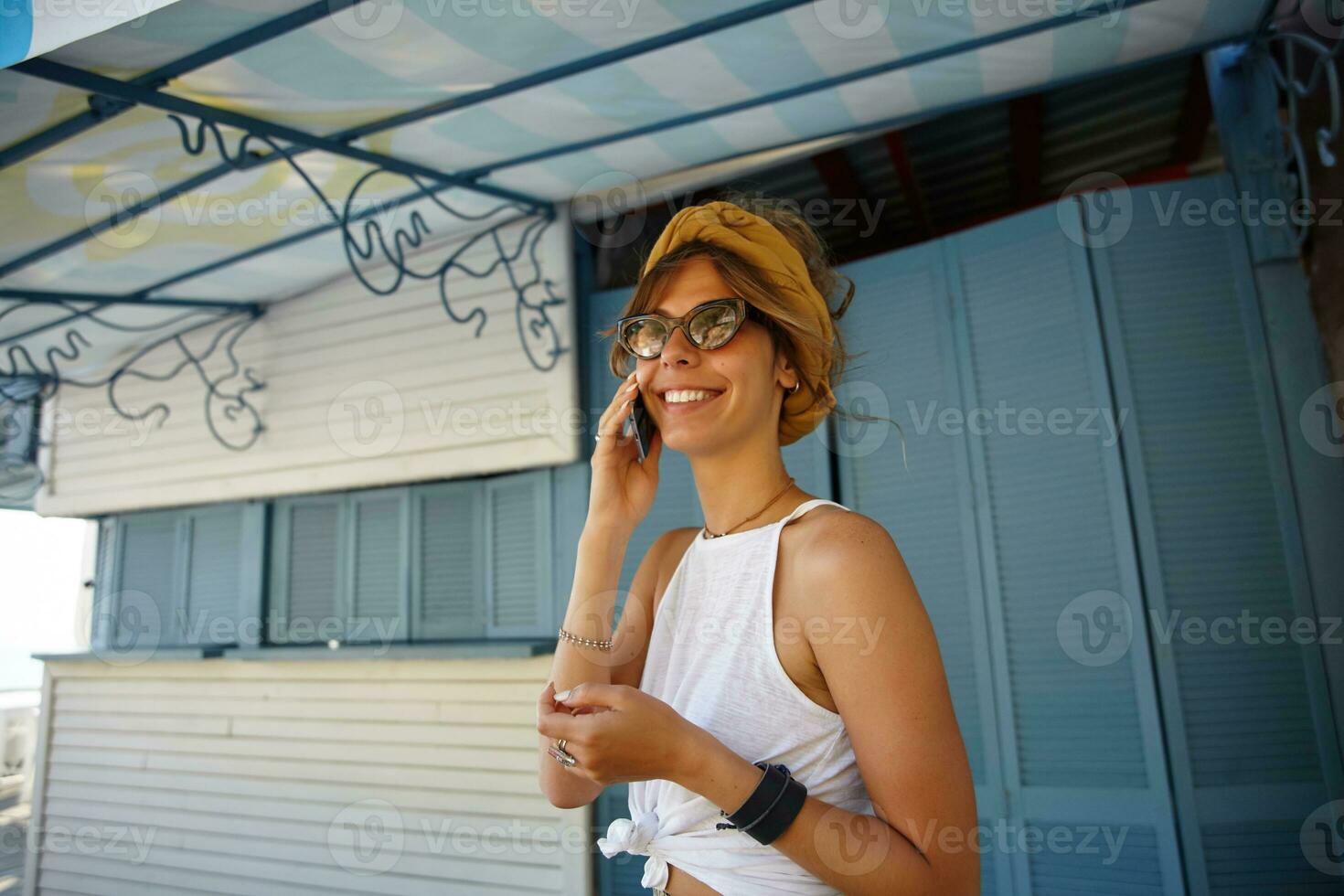 vrolijk jong brunette vrouw in wit top en mosterd hoofdband wandelen buiten Aan warm zonnig dag, maken telefoontje naar vriend en hebben aangenaam praten, wezen in hoog geest foto