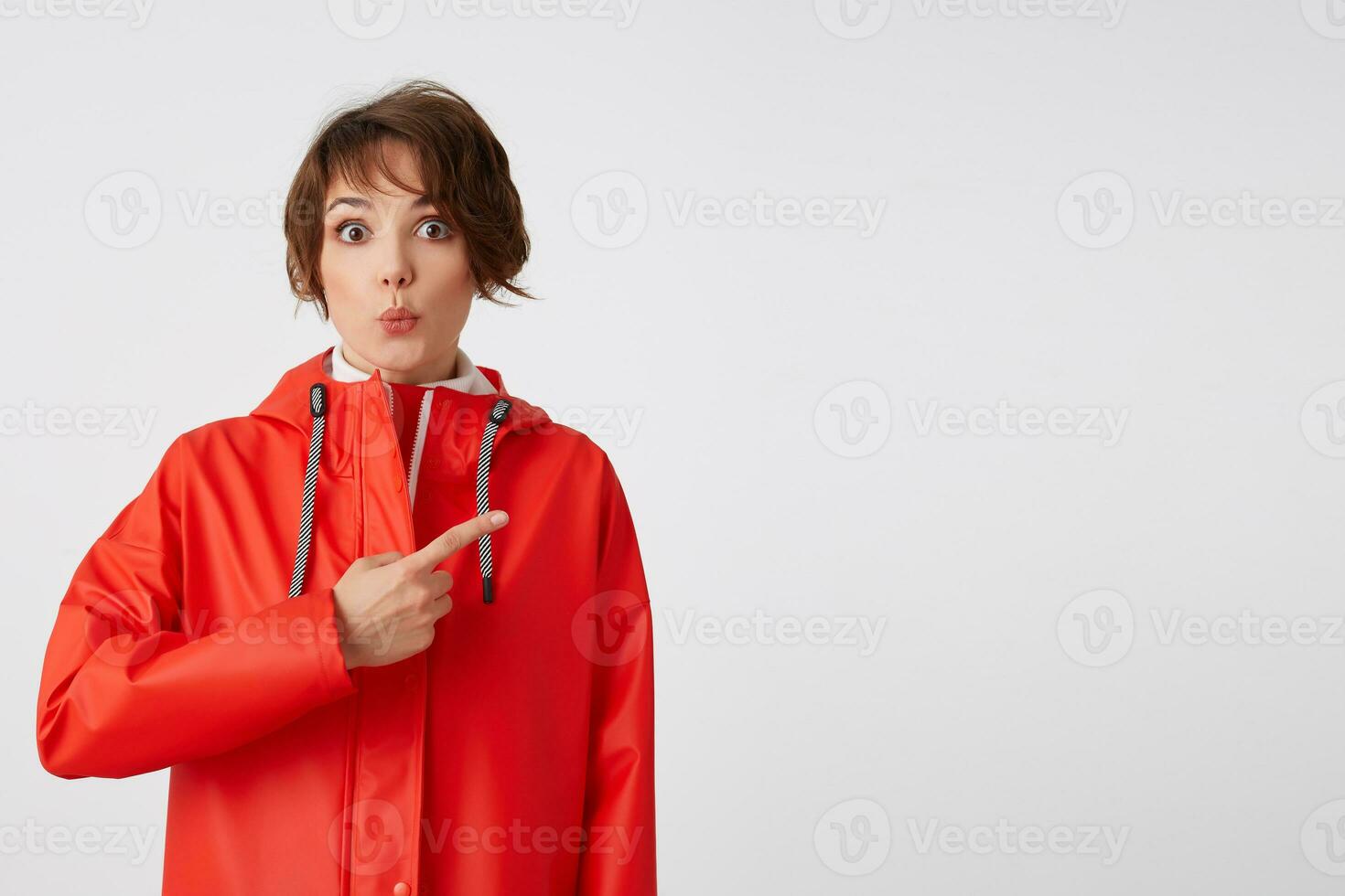 jong schattig gelukkig kort haren vrouw in rood regen jas, hoort koel nieuws. wil naar trek uw aandacht, points vingers naar de kopiëren ruimte. staand over- wit achtergrond. foto