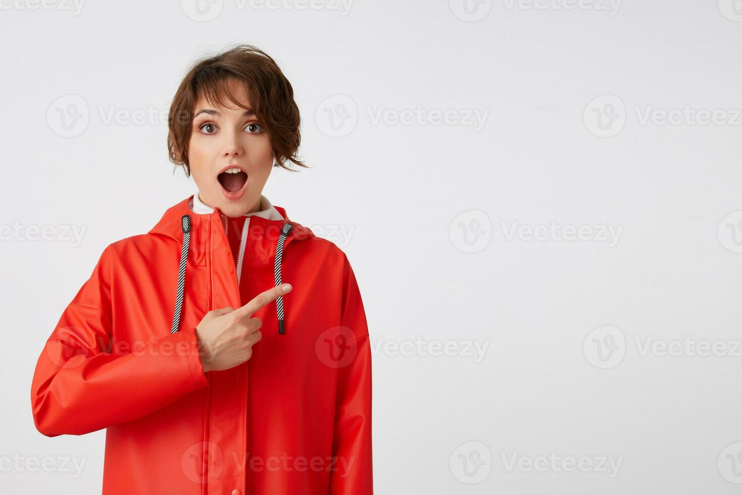 jong mooi hoor gelukkig versteld staan kort haren dame in rood regen jas, met breed Open mond, hoort koel nieuws. wil naar trek uw aandacht, points vingers naar de kopiëren ruimte. staand over- wit achtergrond. foto