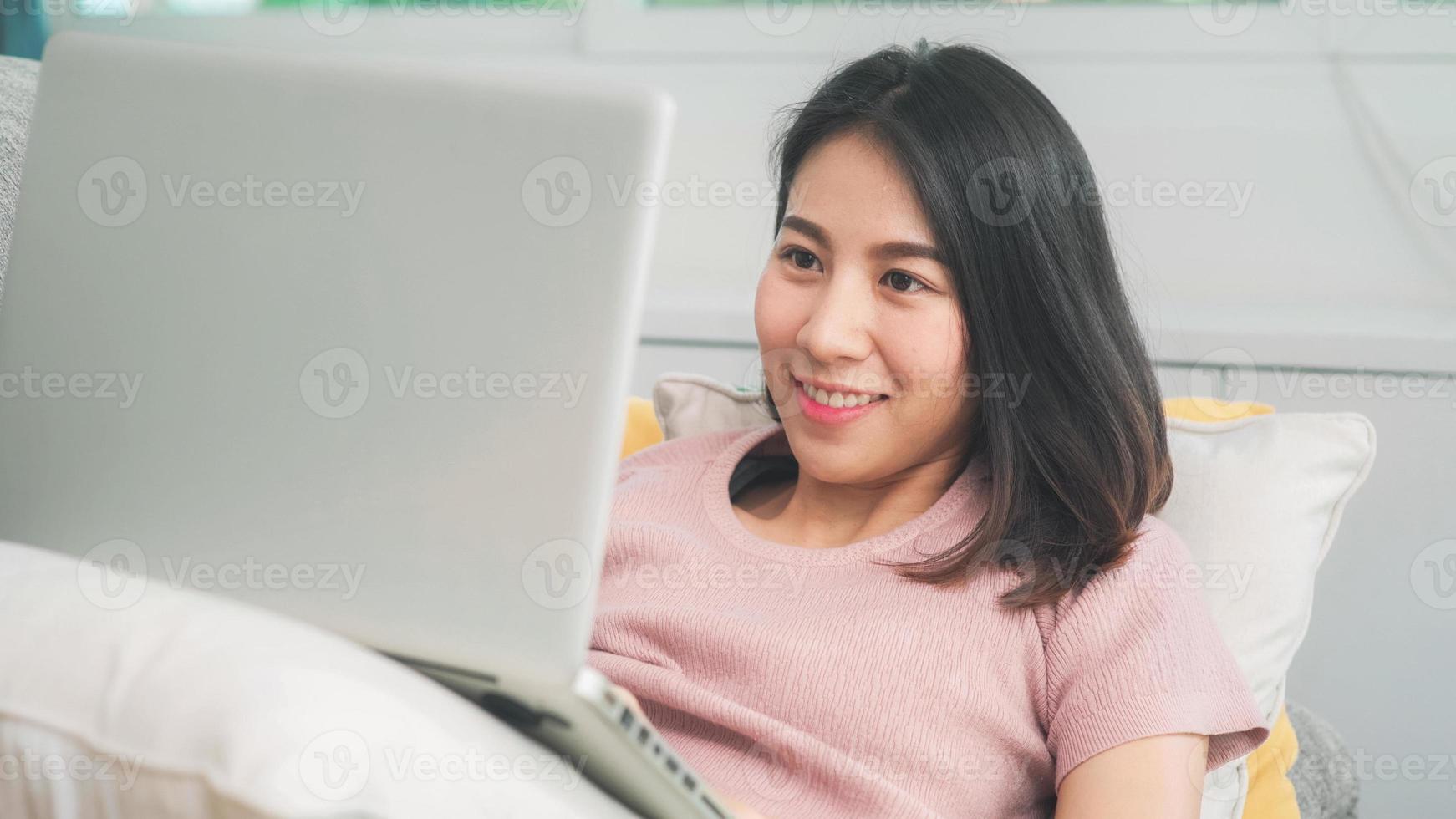 jonge zakelijke freelance aziatische vrouw die op laptop werkt en sociale media controleert terwijl ze op de bank ligt wanneer ze thuis in de woonkamer ontspant. levensstijl Latijnse en Spaanse etniciteit vrouwen bij huis concept. foto