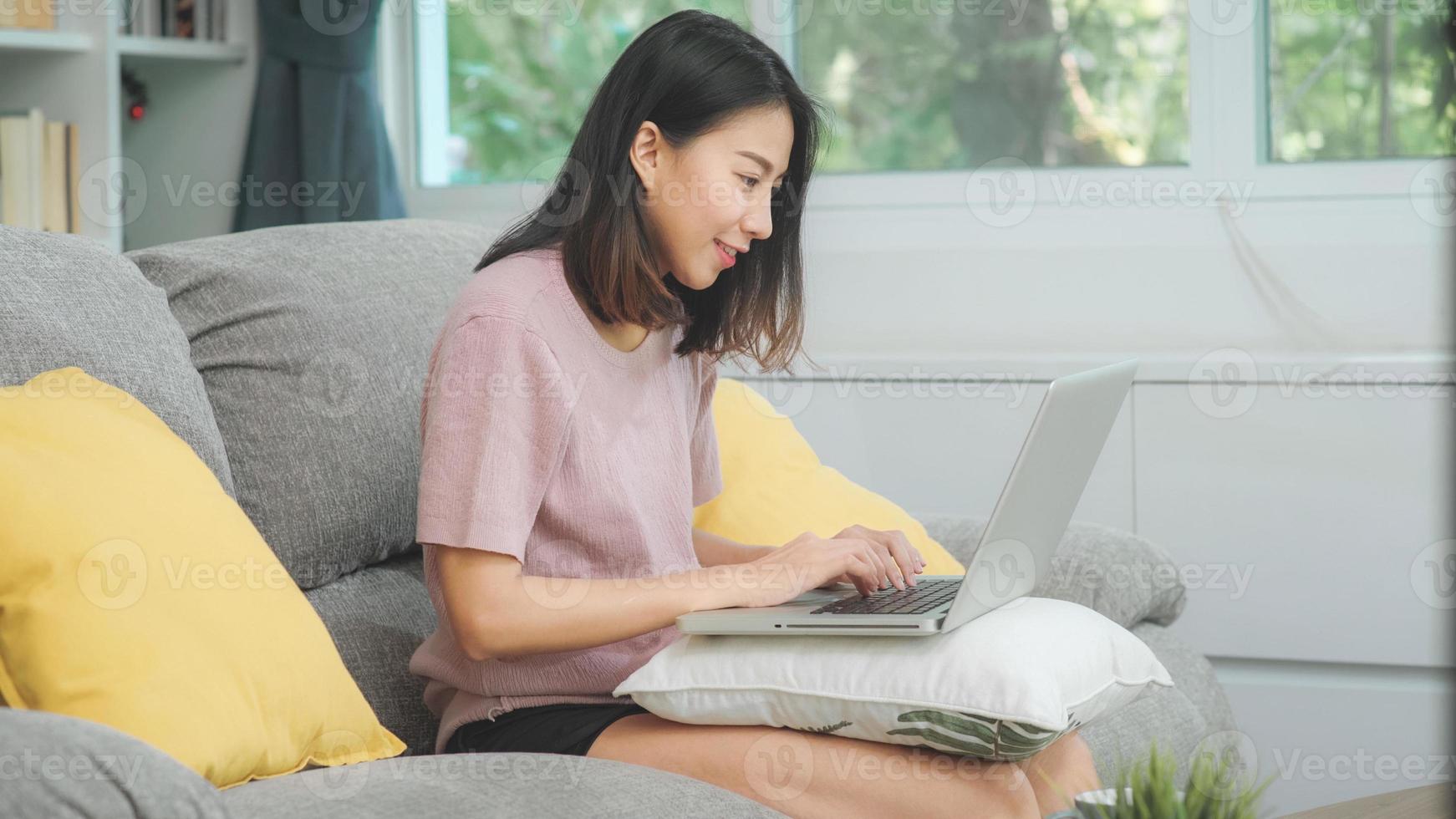 jonge zakelijke freelance aziatische vrouw die op laptop werkt en sociale media controleert terwijl ze op de bank ligt wanneer ze thuis in de woonkamer ontspant. levensstijl Latijnse en Spaanse etniciteit vrouwen bij huis concept. foto