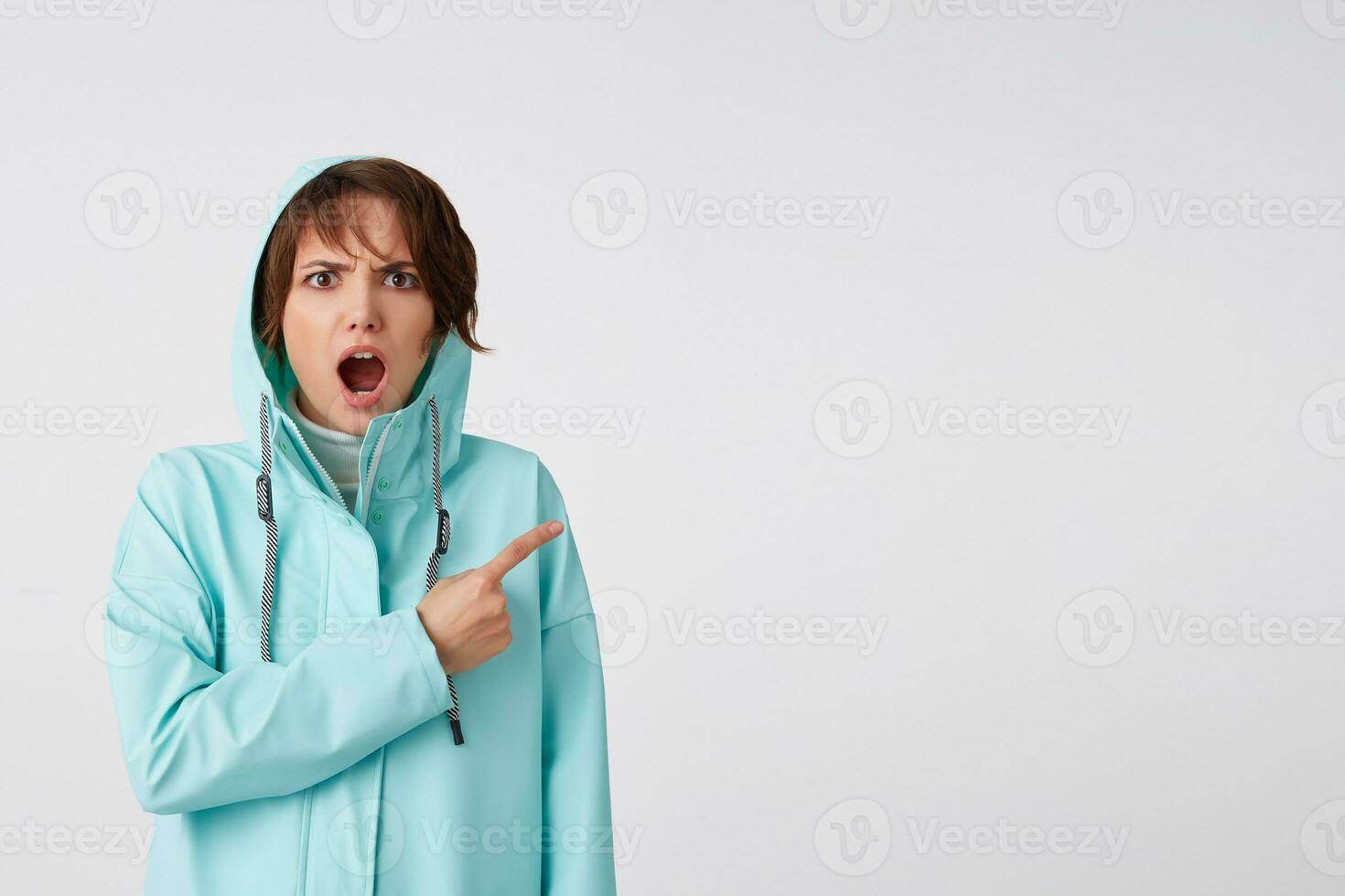geschokt kortharig jong vrouw in blauw regen jas, met breed Open mond en verontwaardigd uitdrukking, wil naar trek u aandacht naar de kopiëren ruimte Aan de Rechtsaf kant, staat over- wit achtergrond. foto