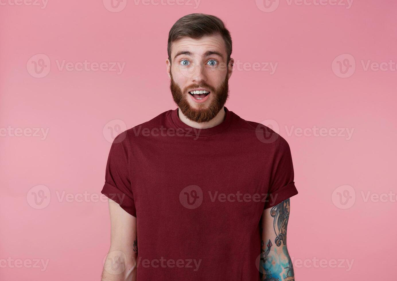 portret van jong knap gelukkig versteld staan rood gebaard Mens in rood t-shirt, standaard over- roze achtergrond, looks Bij de camera met breed Open mond en ogen. foto