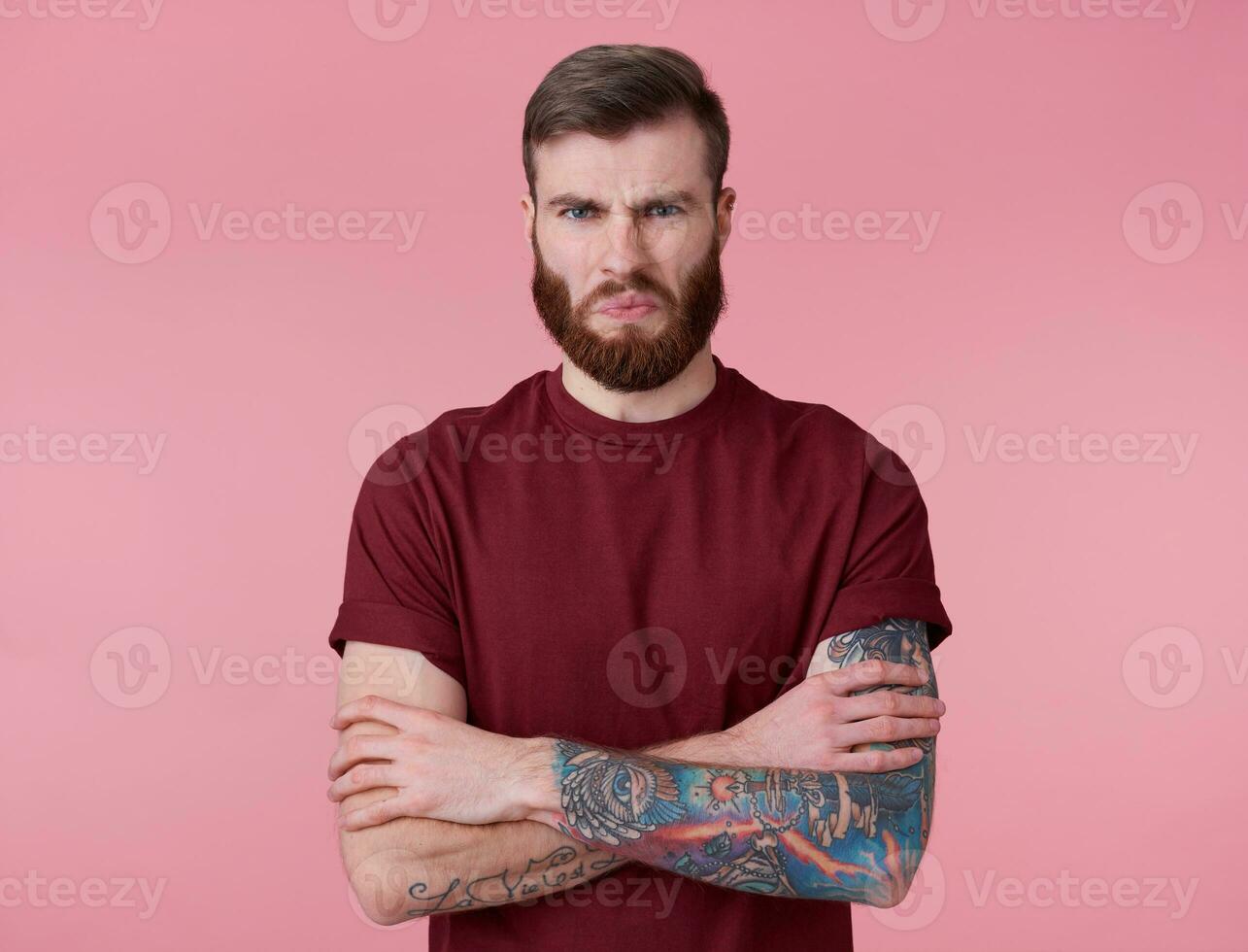 foto van jong knap getatoeëerd walgde rood gebaard Mens in blanco t-shirt, staat met gekruiste armen over- roze achtergrond, fronsen en looks Bij de camera.