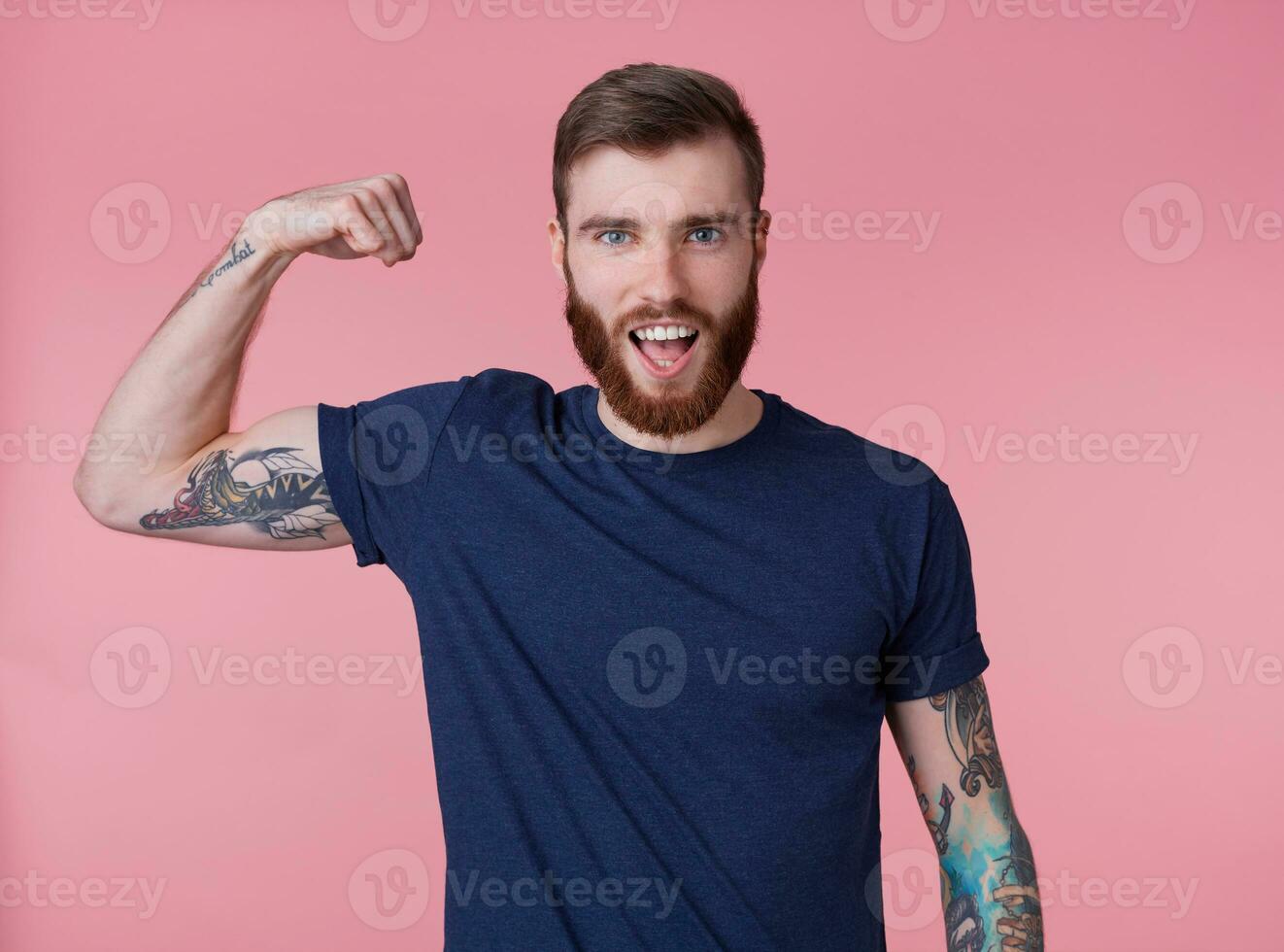 jong roodbaard knap manvol vent demonstreert biceps en sterkte met breed Open mond, op zoek Bij de camera geïsoleerd over- roze achtergrond. foto